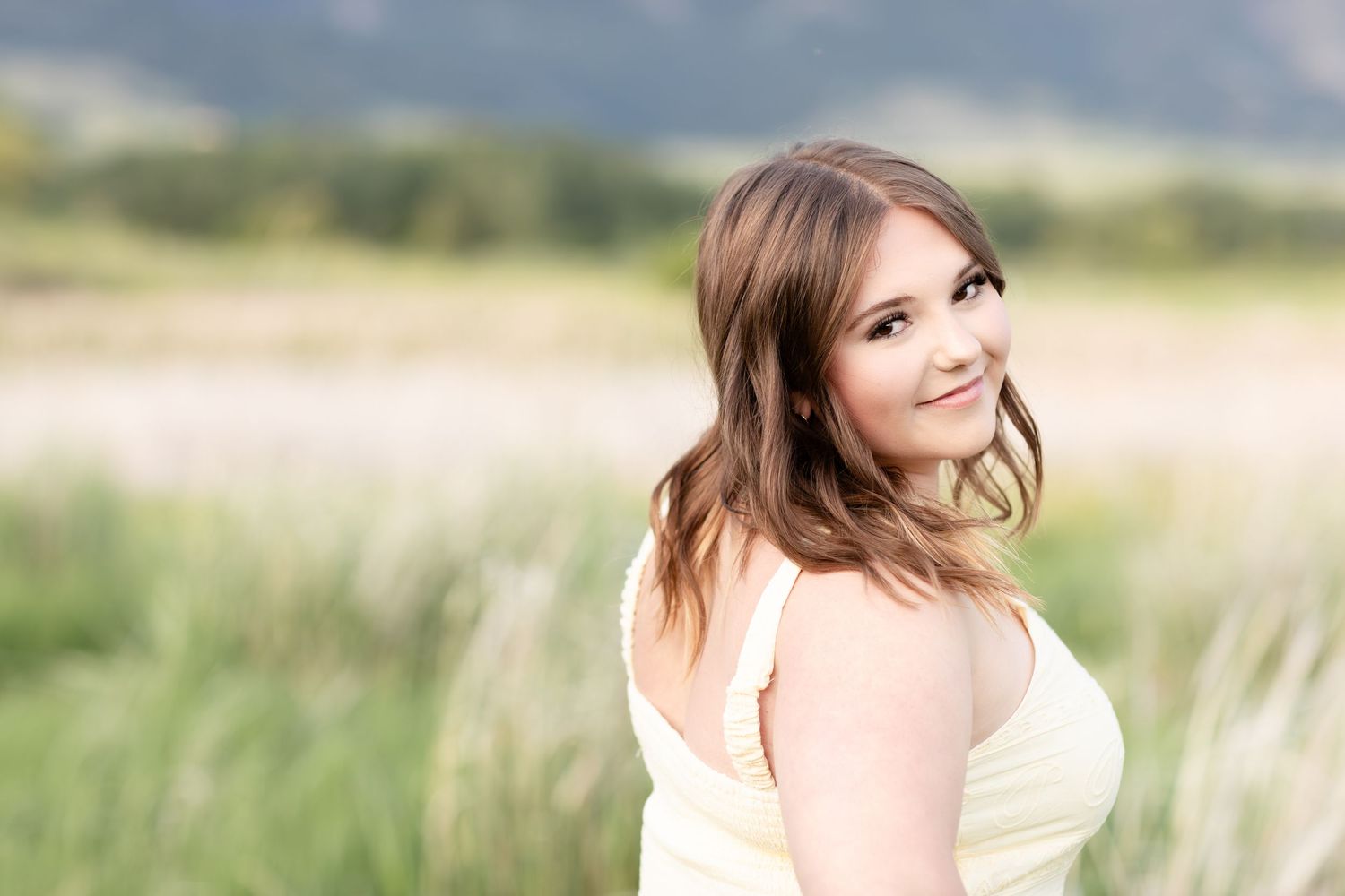 Young woman in white dress smiling over shoulder in sunlit meadow with mountains in background.