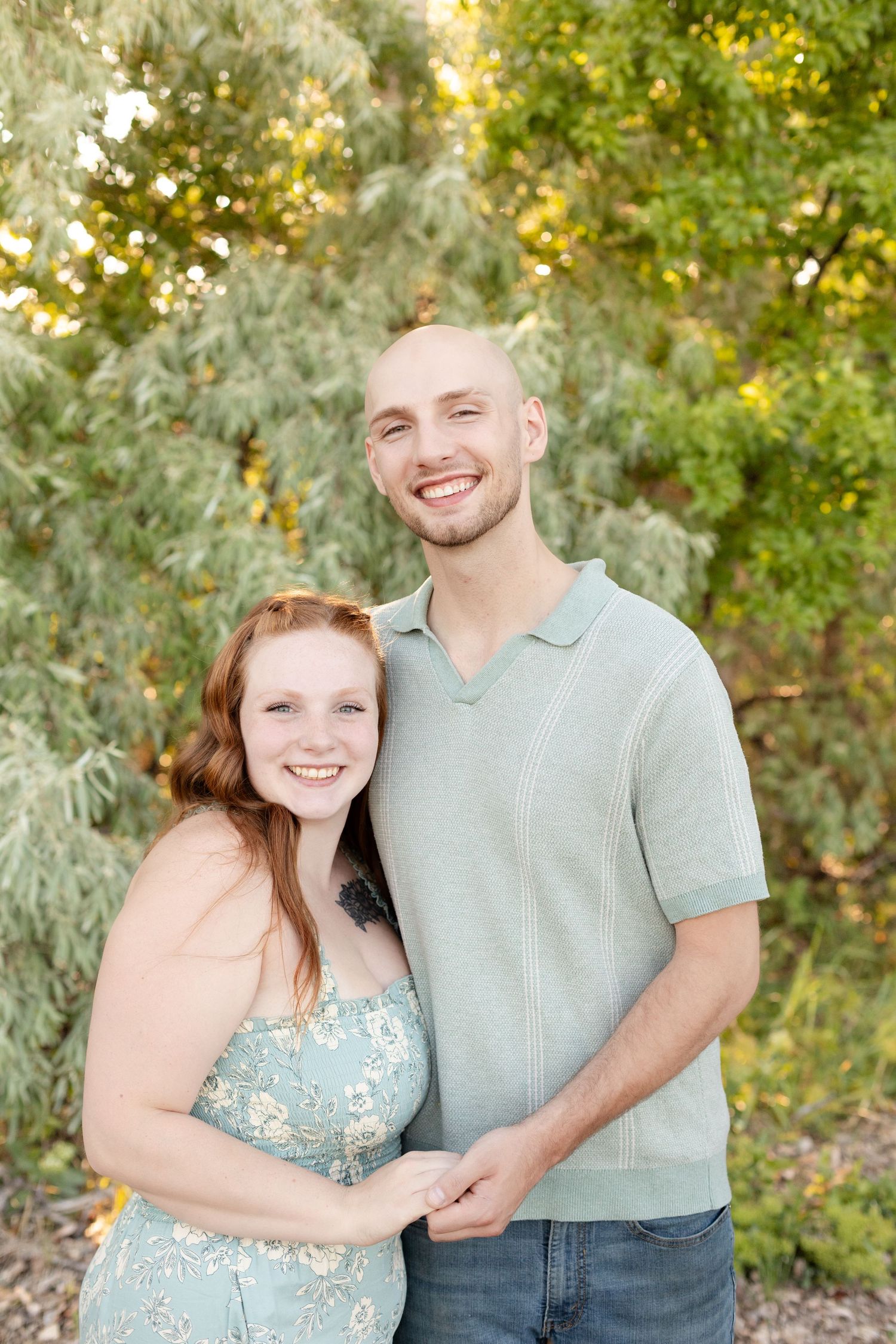 Happy couple holding hands and smiling in front of lush green trees outdoors.