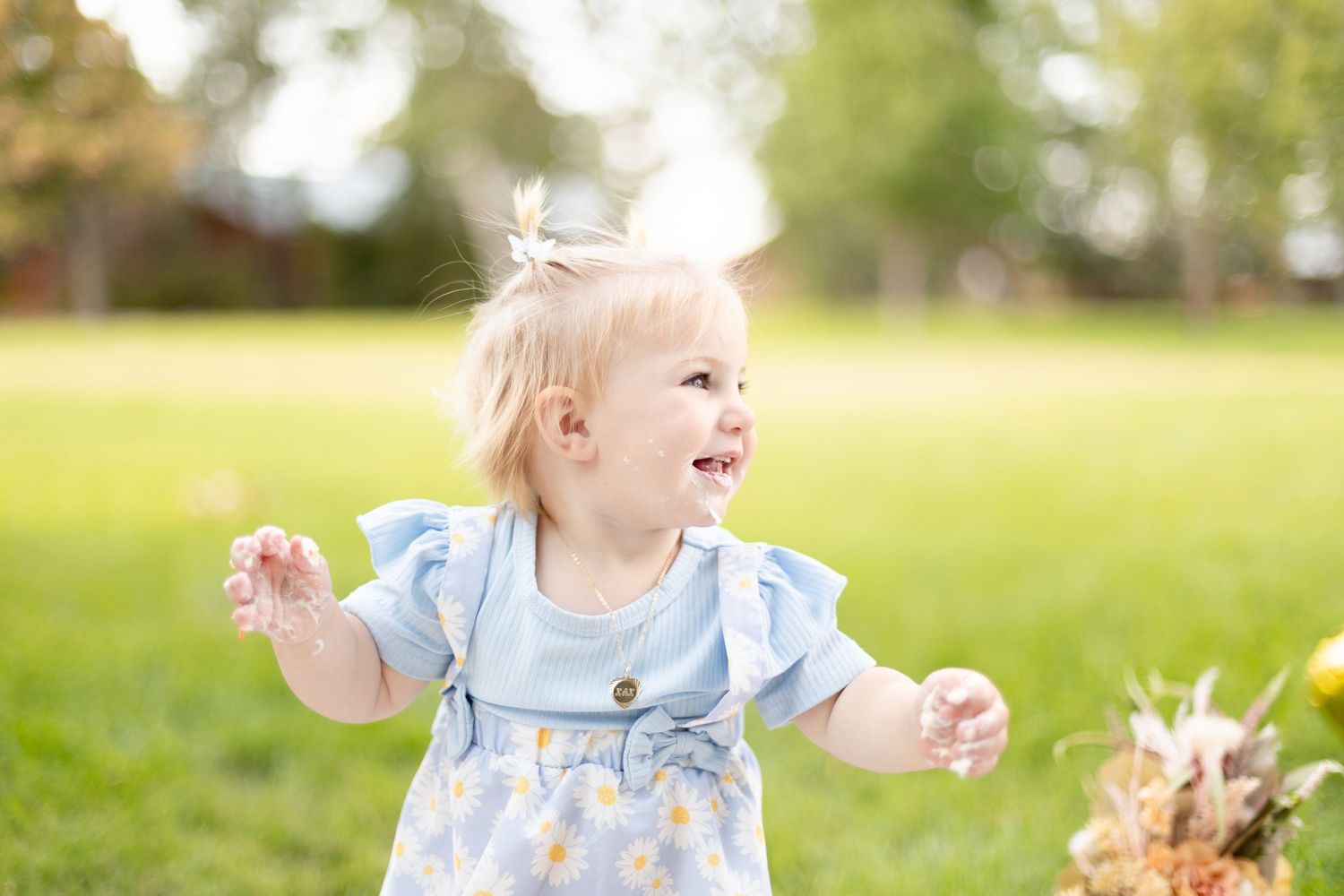 Smiling blonde toddler girl in blue daisy dress playing outdoors in green grass park.