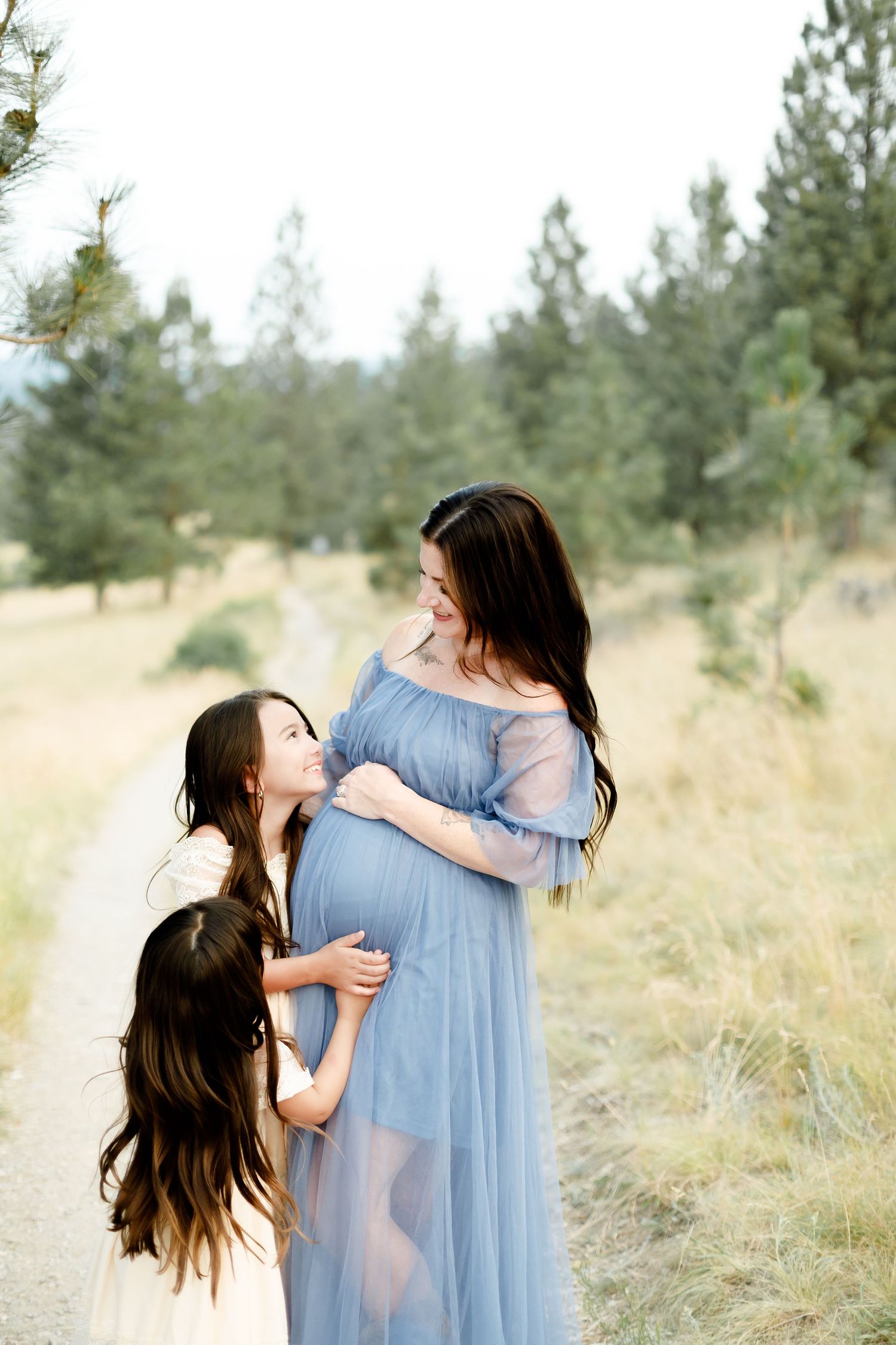 Pregnant woman in blue maternity gown embraced by dark-haired daughter on forest path.