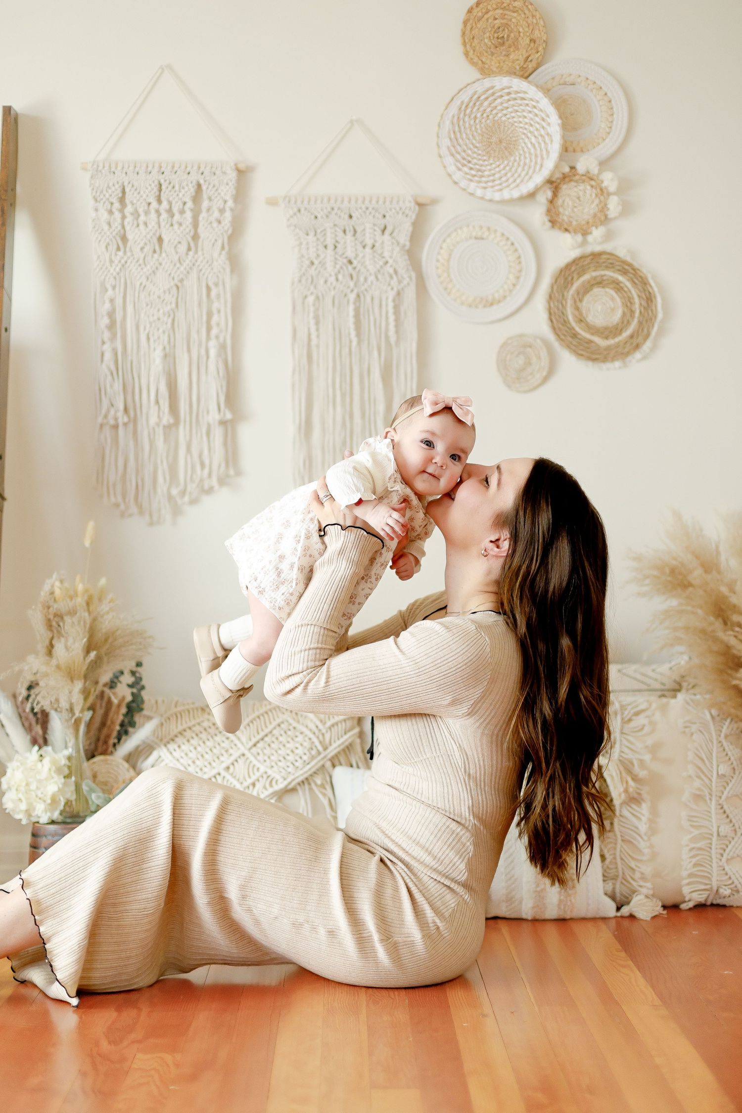 Mother in cream dress sitting on floor kissing baby girl lifted in air with boho decor backdrop.