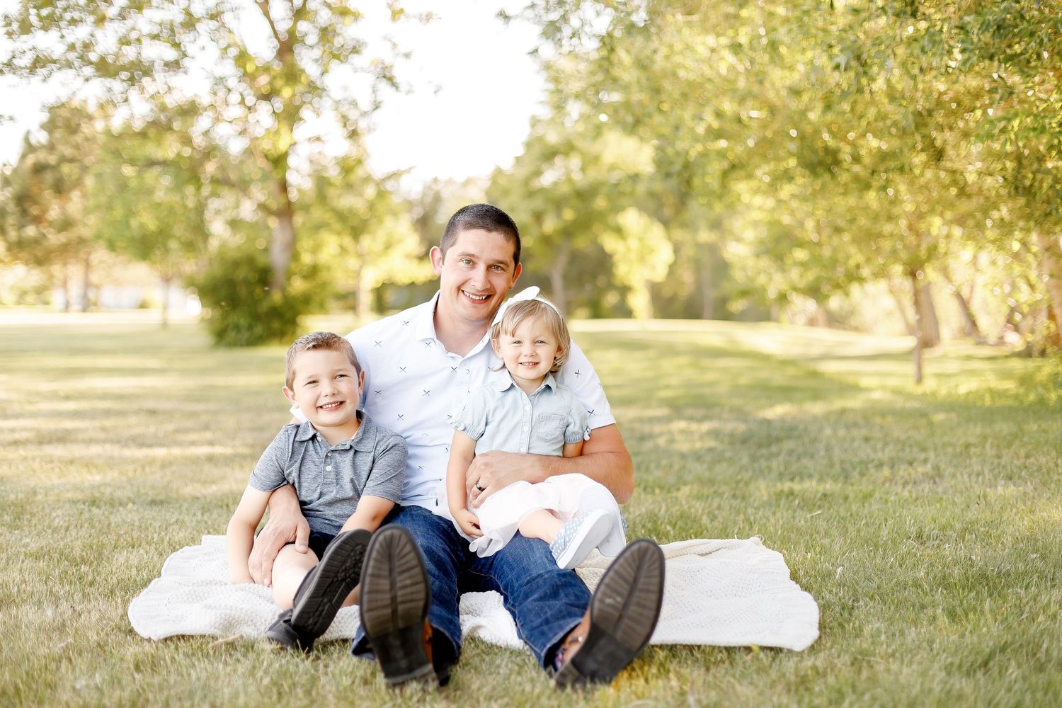 Father sits on a blanket in a park with his two young sons smiling.