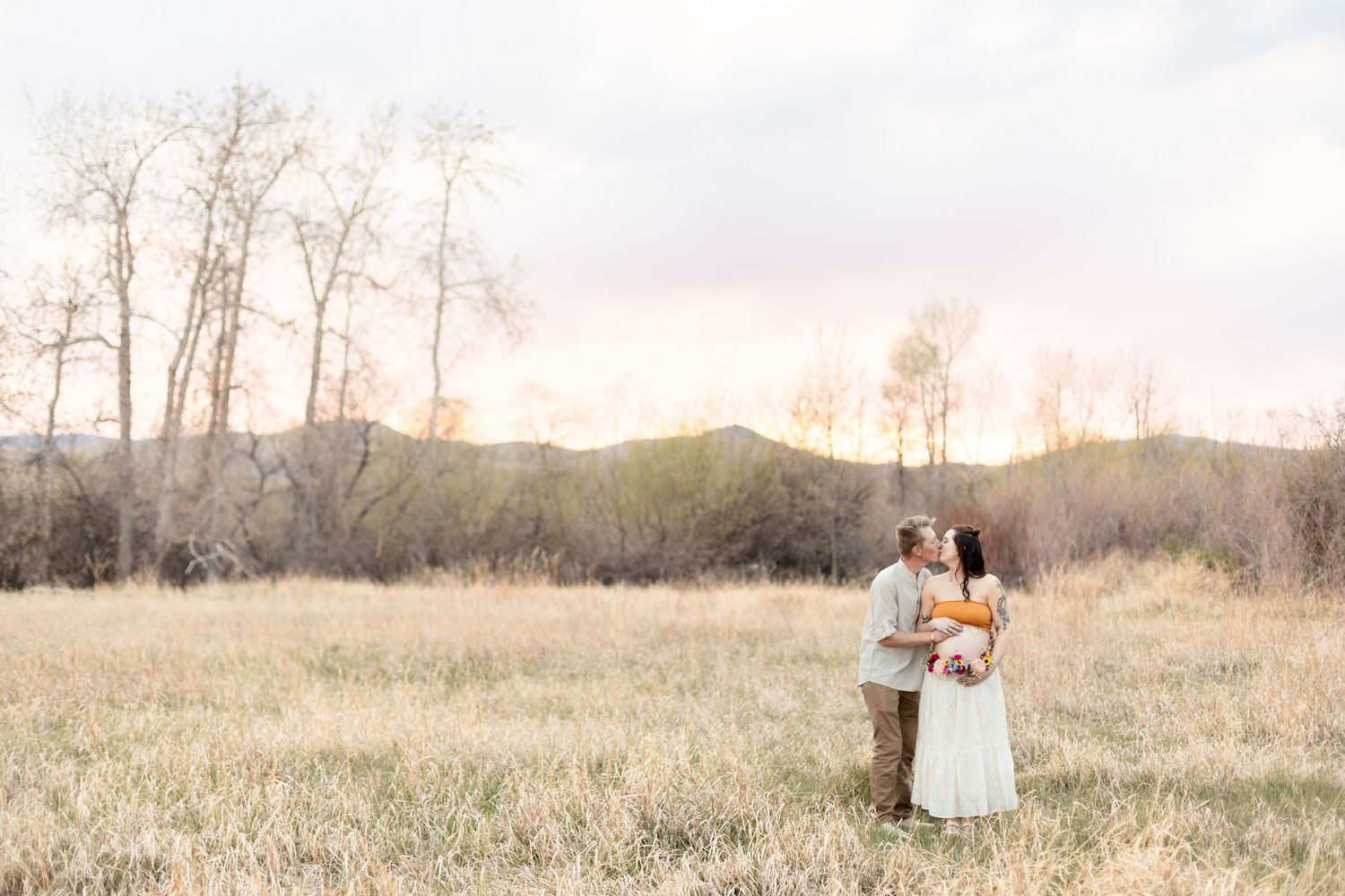 Couple sharing a tender kiss in a sunlit meadow with bare trees and mountains in the background.