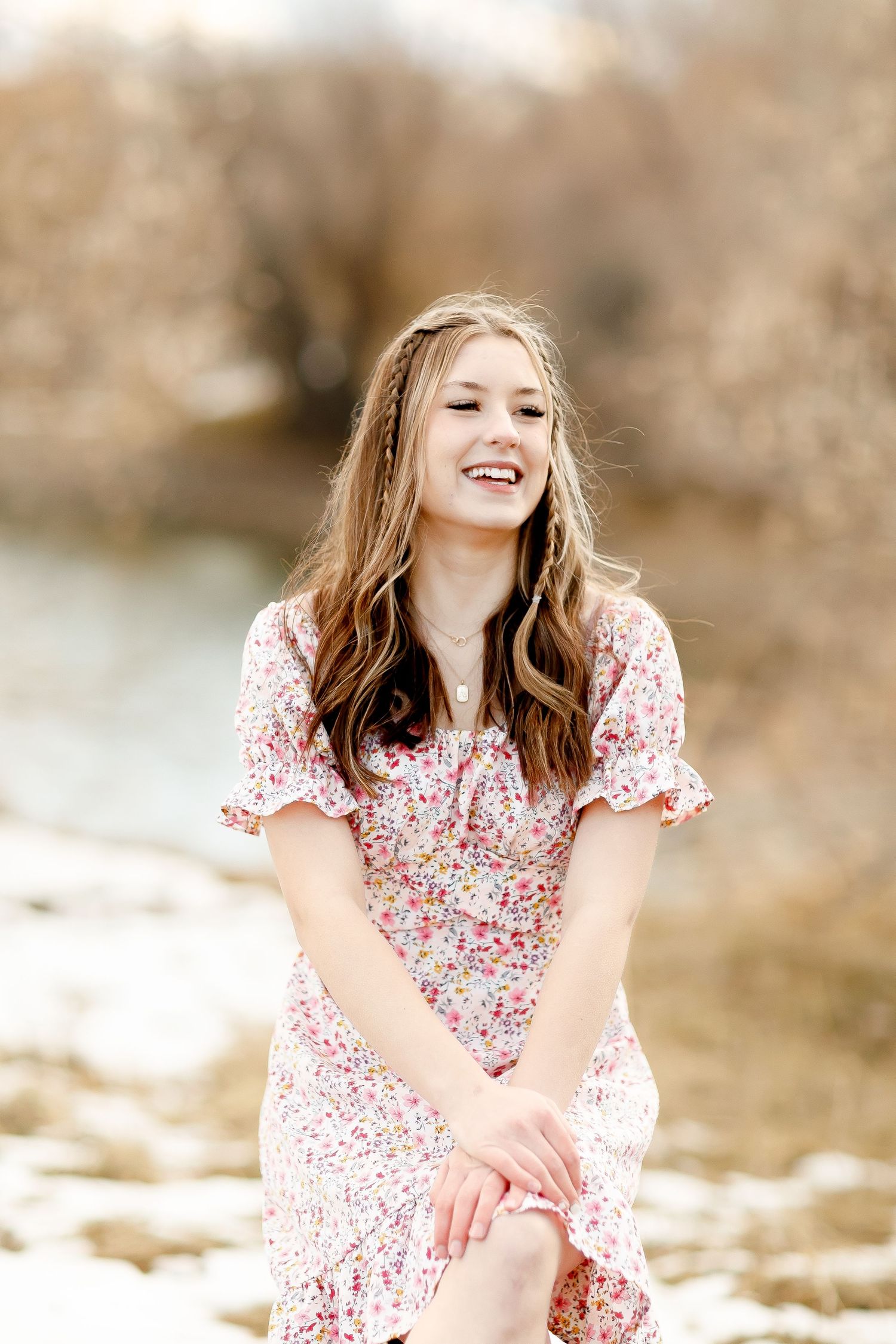 Laughing teenage girl in floral dress sitting outdoors near a snowy riverbank in winter.