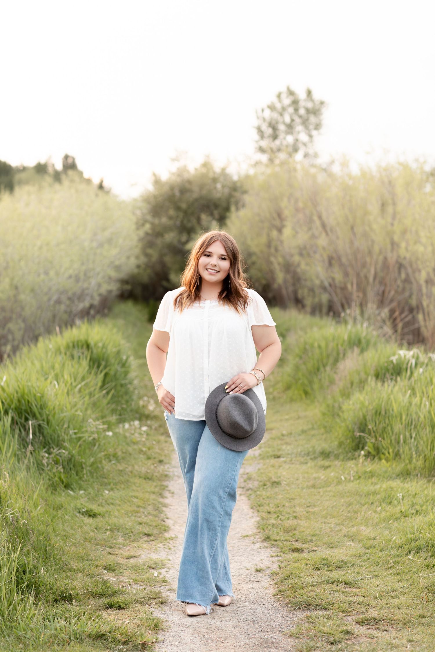 A smiling young woman in a white top and flare jeans holds a hat on a green nature path.