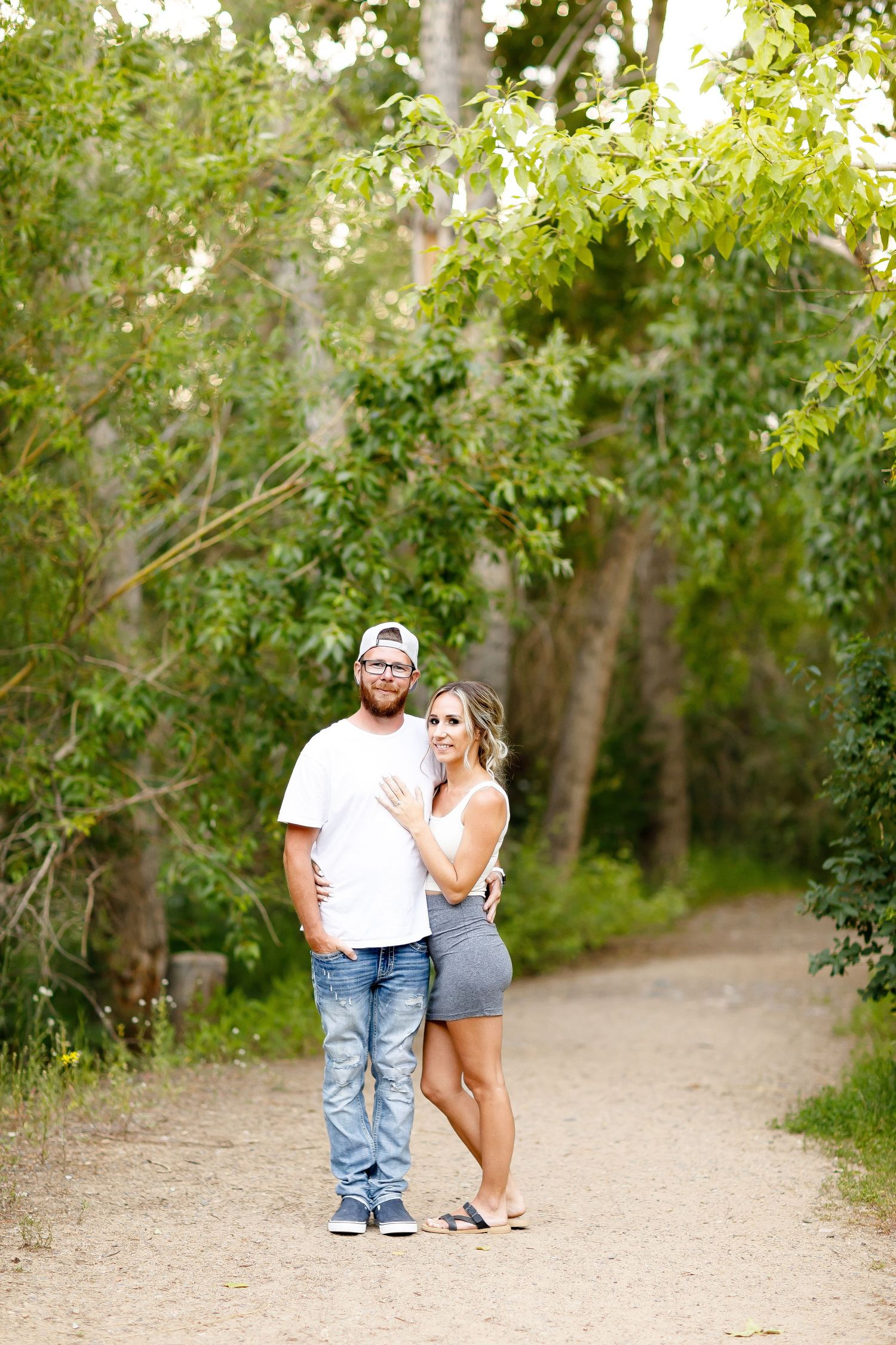 Engaged couple posing together on a shaded garden path surrounded by lush green trees.