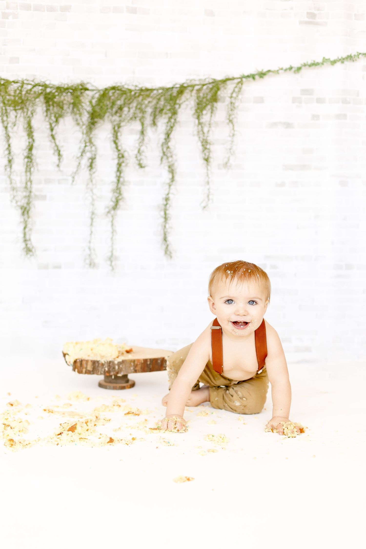 A smiling baby boy in suspenders crawling away from a smashed birthday cake on a white backdrop.