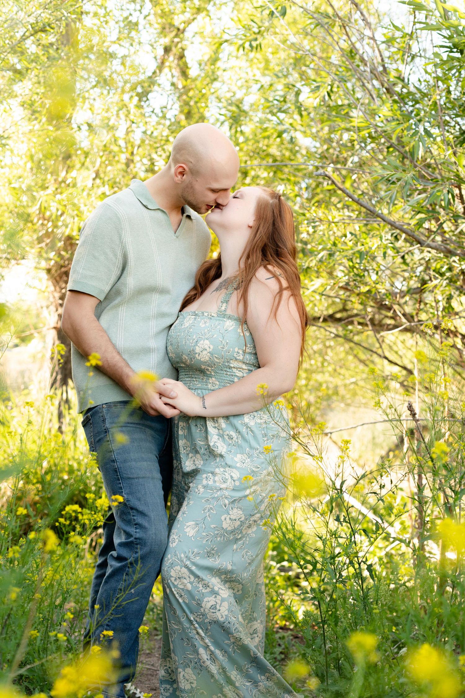 Couple sharing a kiss surrounded by yellow wildflowers and sunlit green trees outdoors.