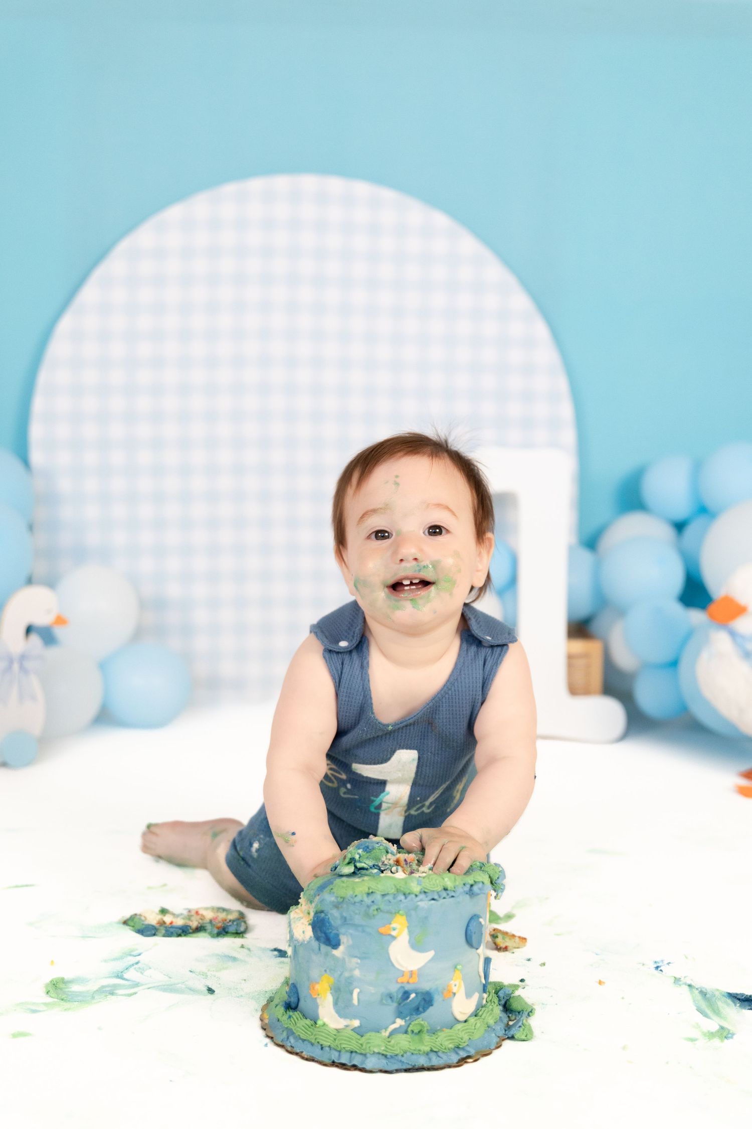 Smiling baby boy with green frosting on face sits beside smashed blue duck first birthday cake.