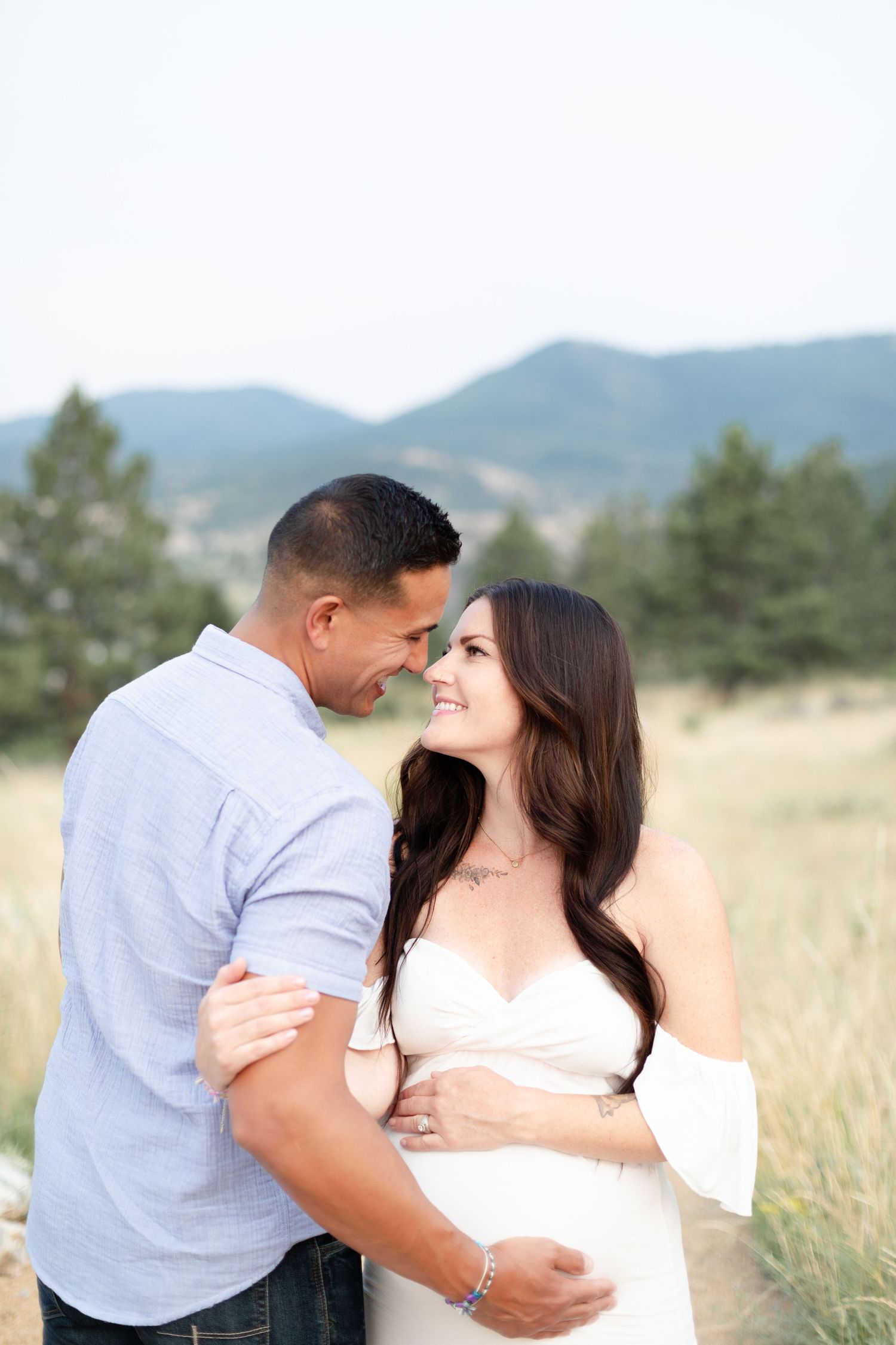 Smiling pregnant woman in white off-shoulder gown embraces partner in a mountain meadow setting.