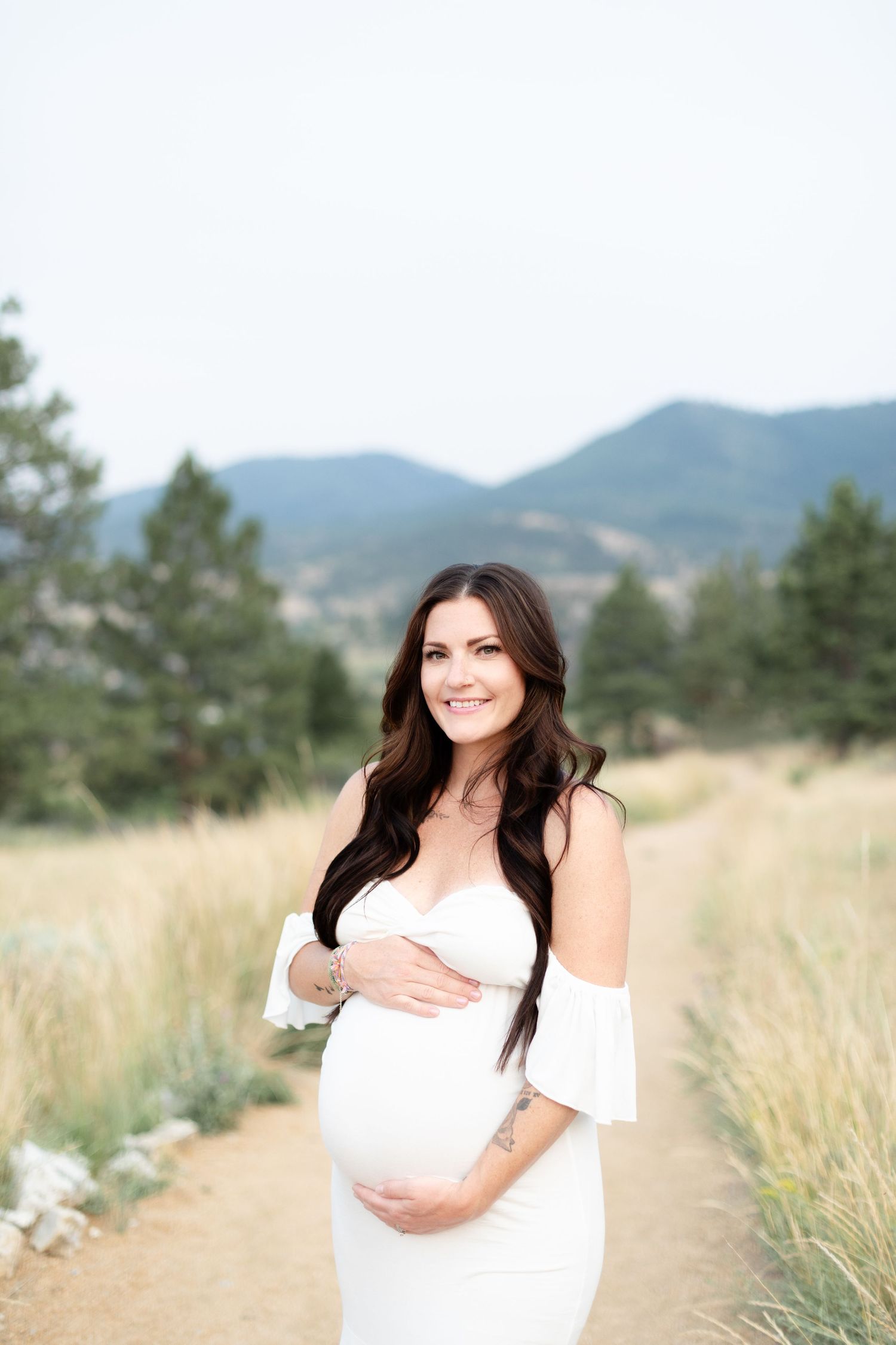 Radiant pregnant woman in white maternity gown posing on scenic mountain trail.