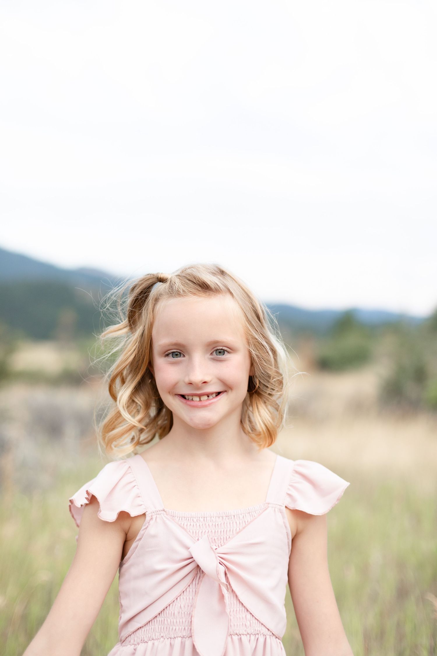 Cheerful young blonde girl in a pink dress smiling in a sunny mountain meadow.