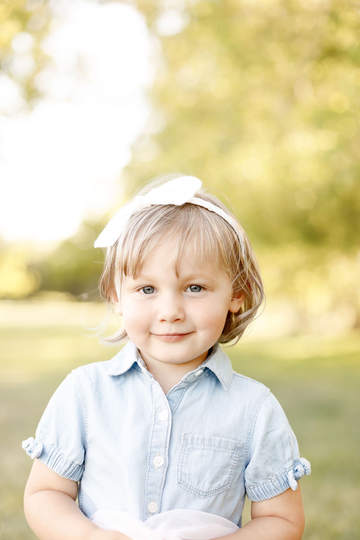 Adorable toddler girl with white bow headband smiles in a sunny park.