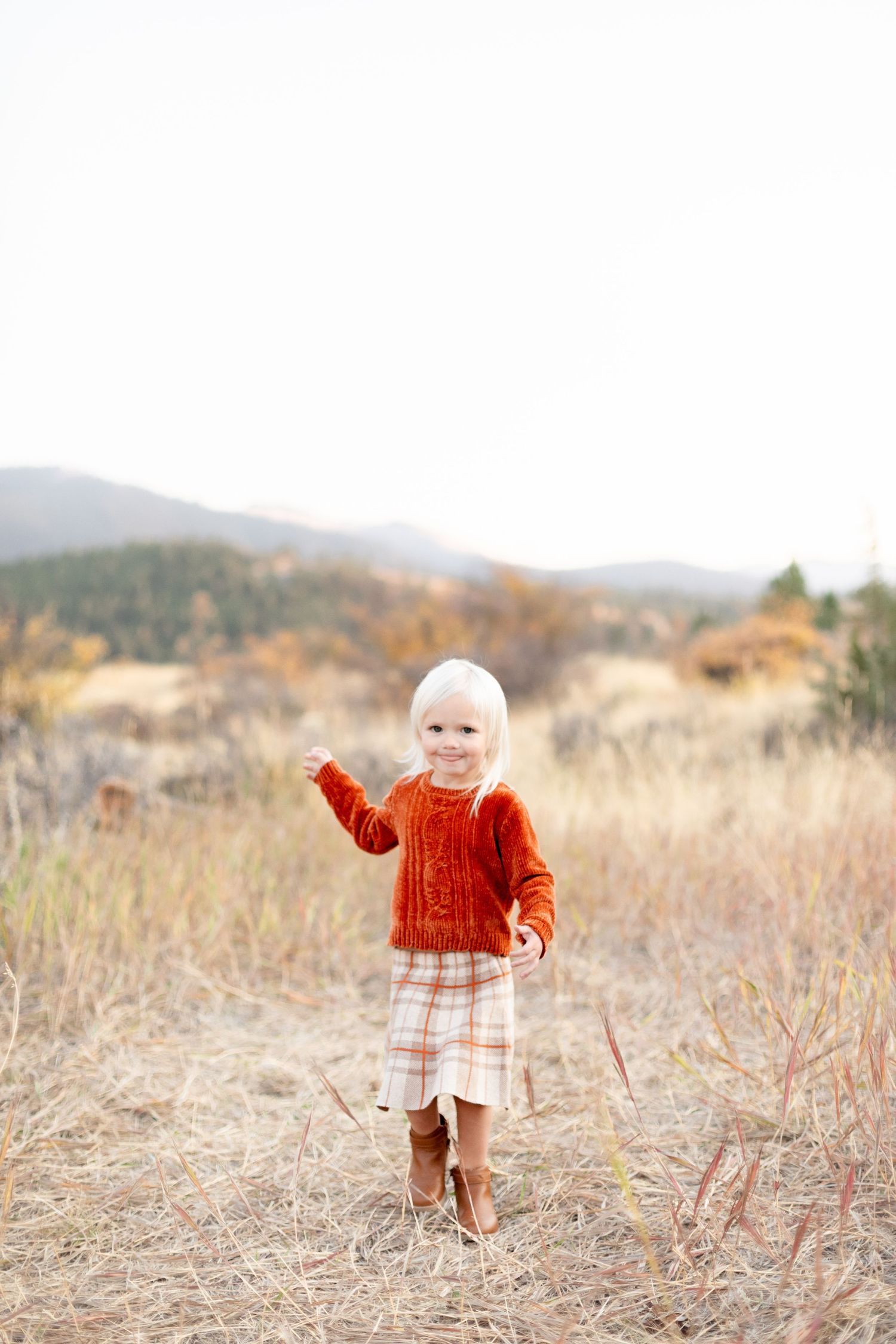 Adorable blonde toddler girl in orange sweater and plaid skirt standing in an autumn meadow.
