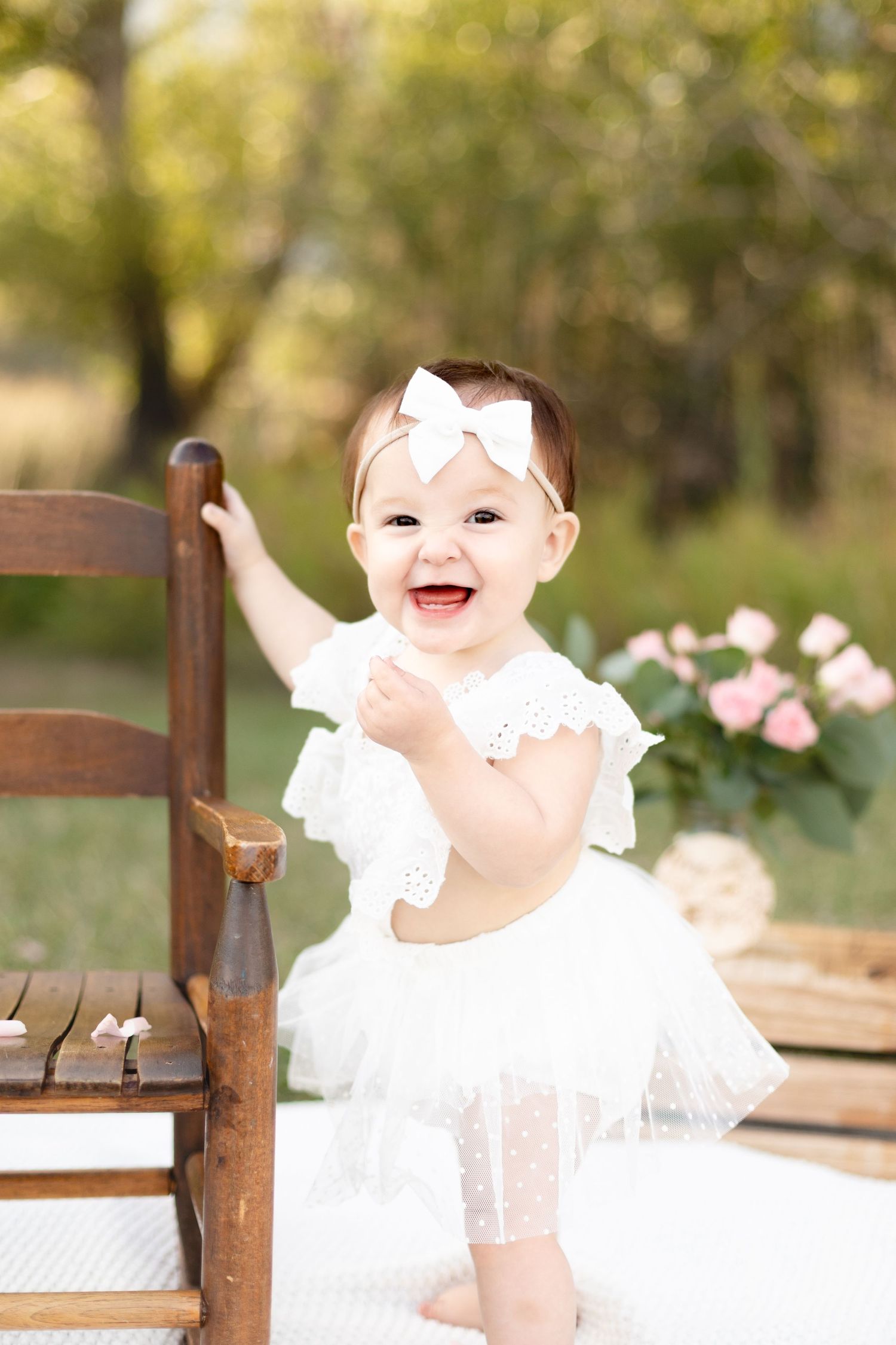 Joyful baby girl in white lace dress standing by wooden rocking chair at outdoor birthday session.