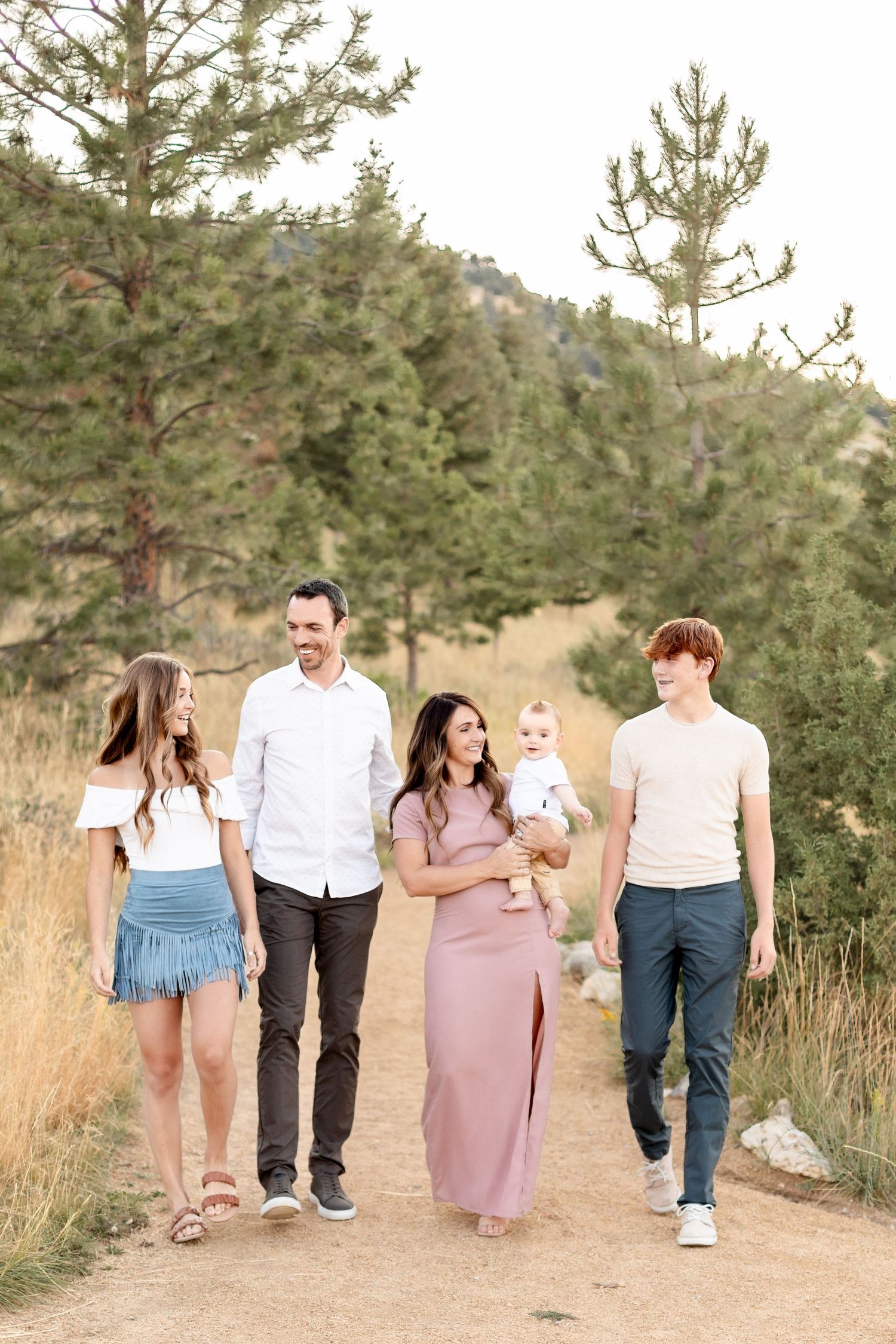 A family of five walks together on a dirt path surrounded by pine trees and golden hills.