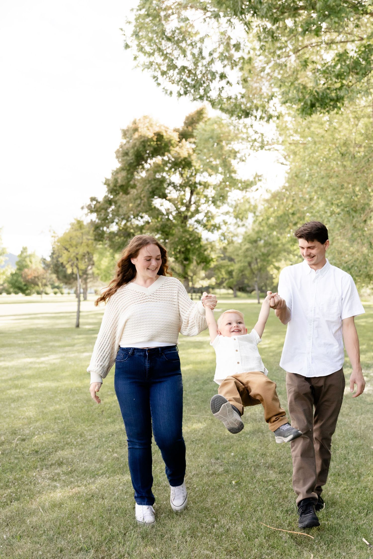 A young couple swings their laughing toddler between them while walking in a sunny green park.