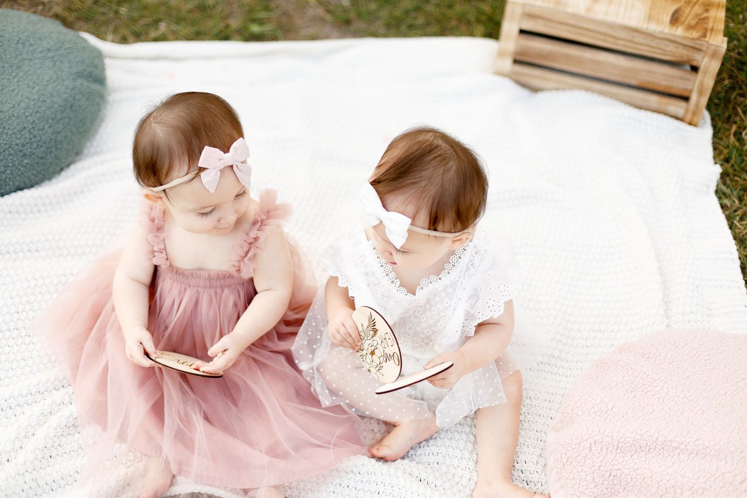 Two baby girls in pink and white dresses sitting together on blanket during outdoor birthday photos.
