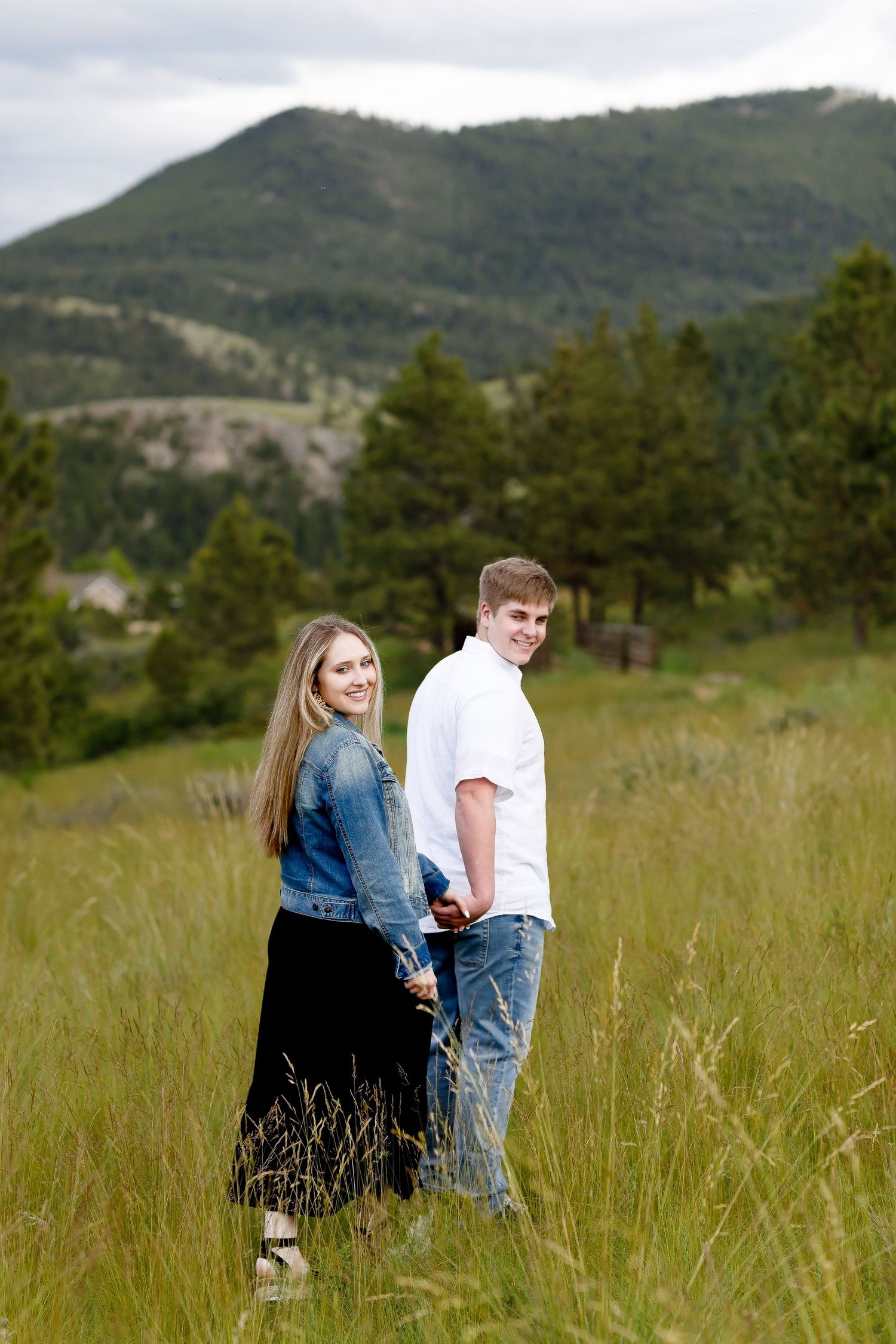 Happy engaged couple holding hands in a mountain meadow with tall grass and pine trees.