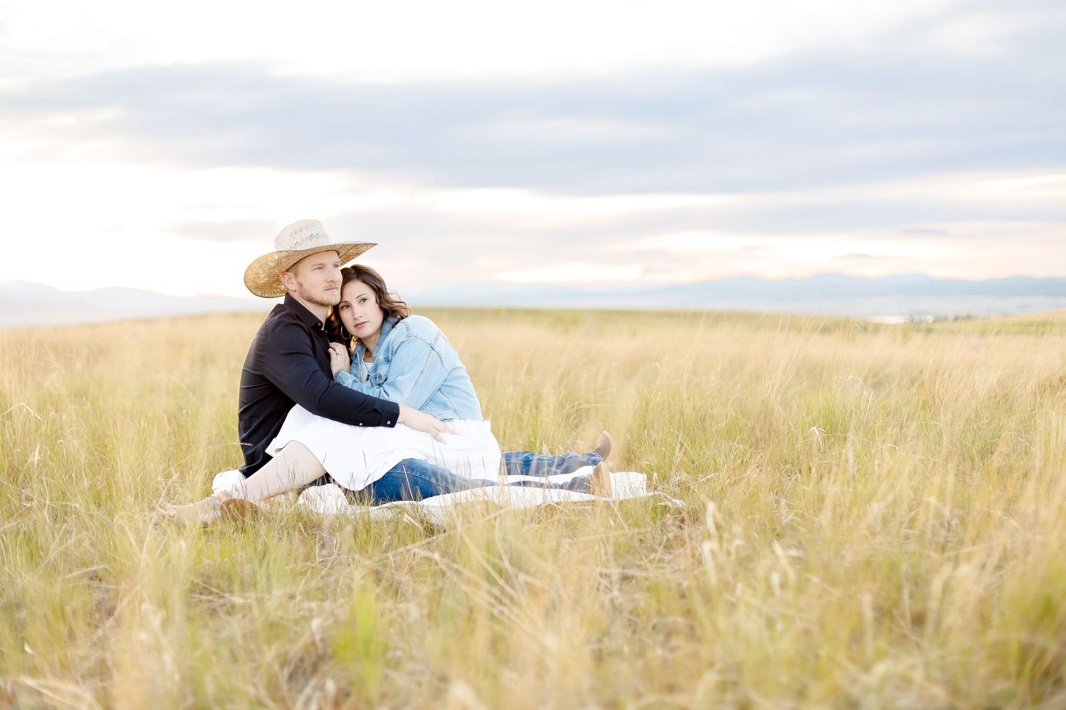 Couple sitting together in tall golden grass field, man in black shirt and cowboy hat, woman in denim jacket.