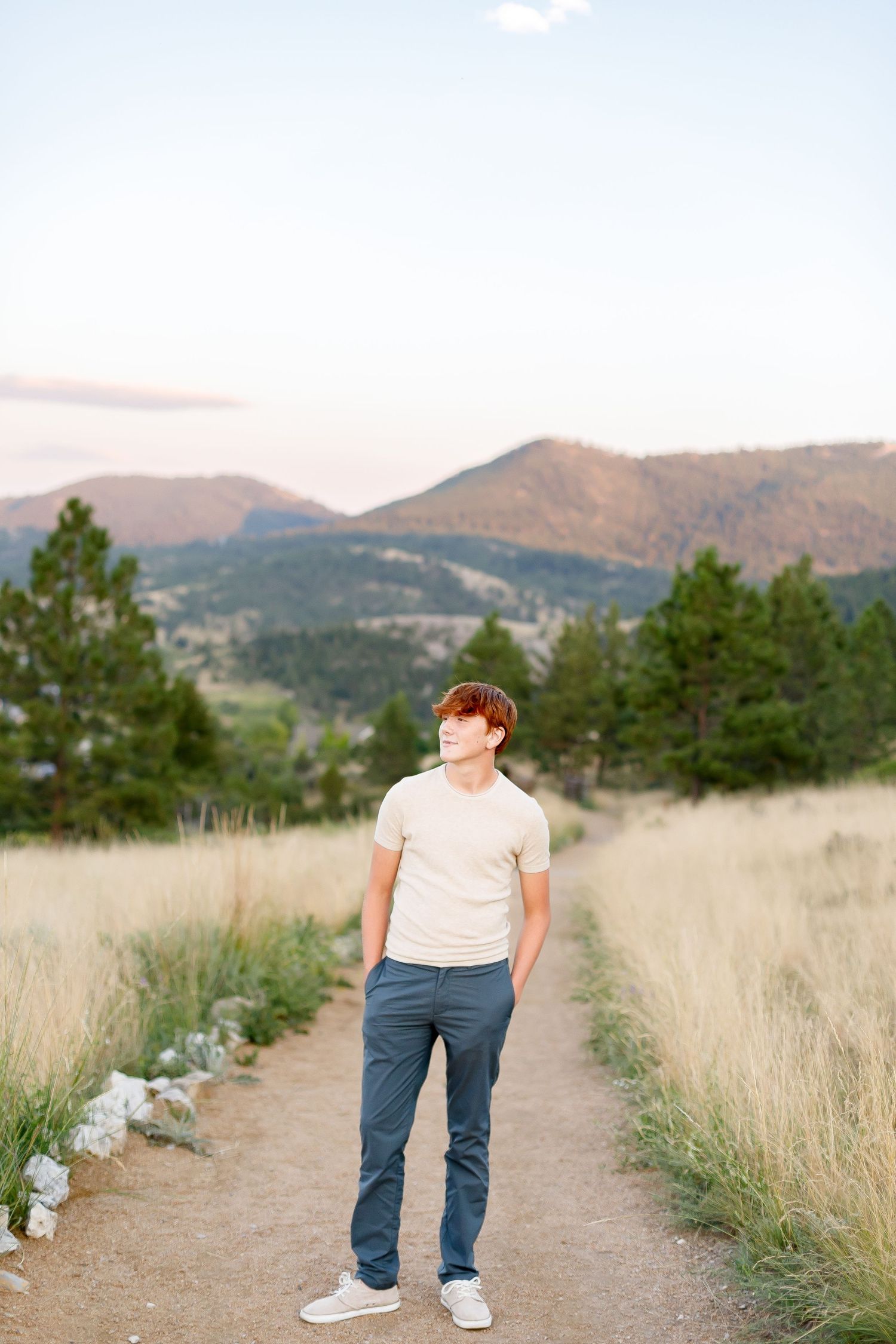 Red-haired teenage boy in beige shirt and gray pants standing on a dirt path with mountain views.