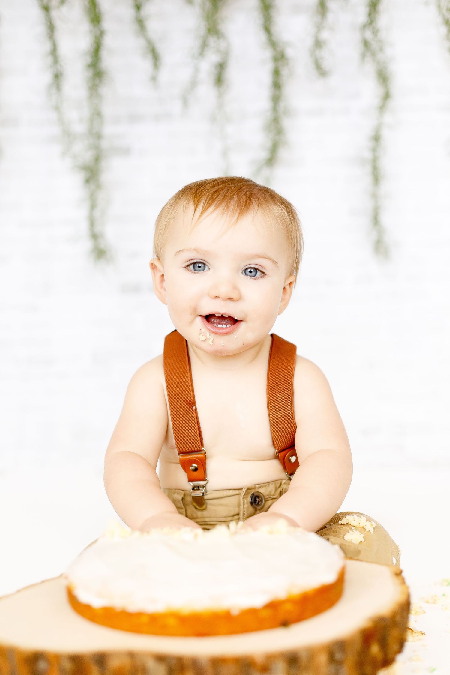 A smiling baby boy with blue eyes wearing suspenders sits behind a white frosted cake on a wooden stump.