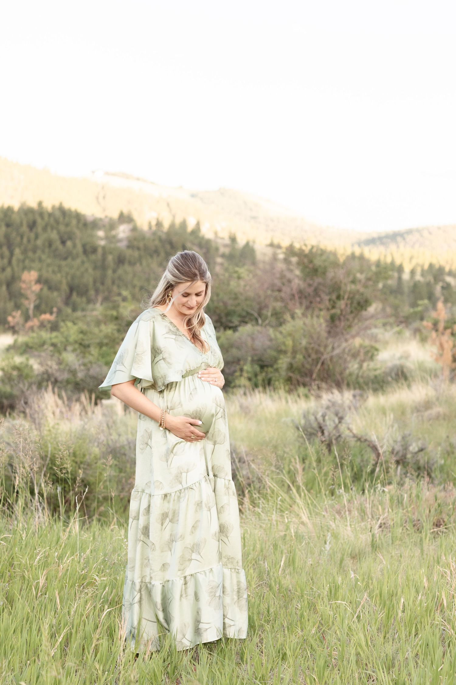 Pregnant woman in sage green floral maxi dress standing in mountain meadow looking down at baby bump.