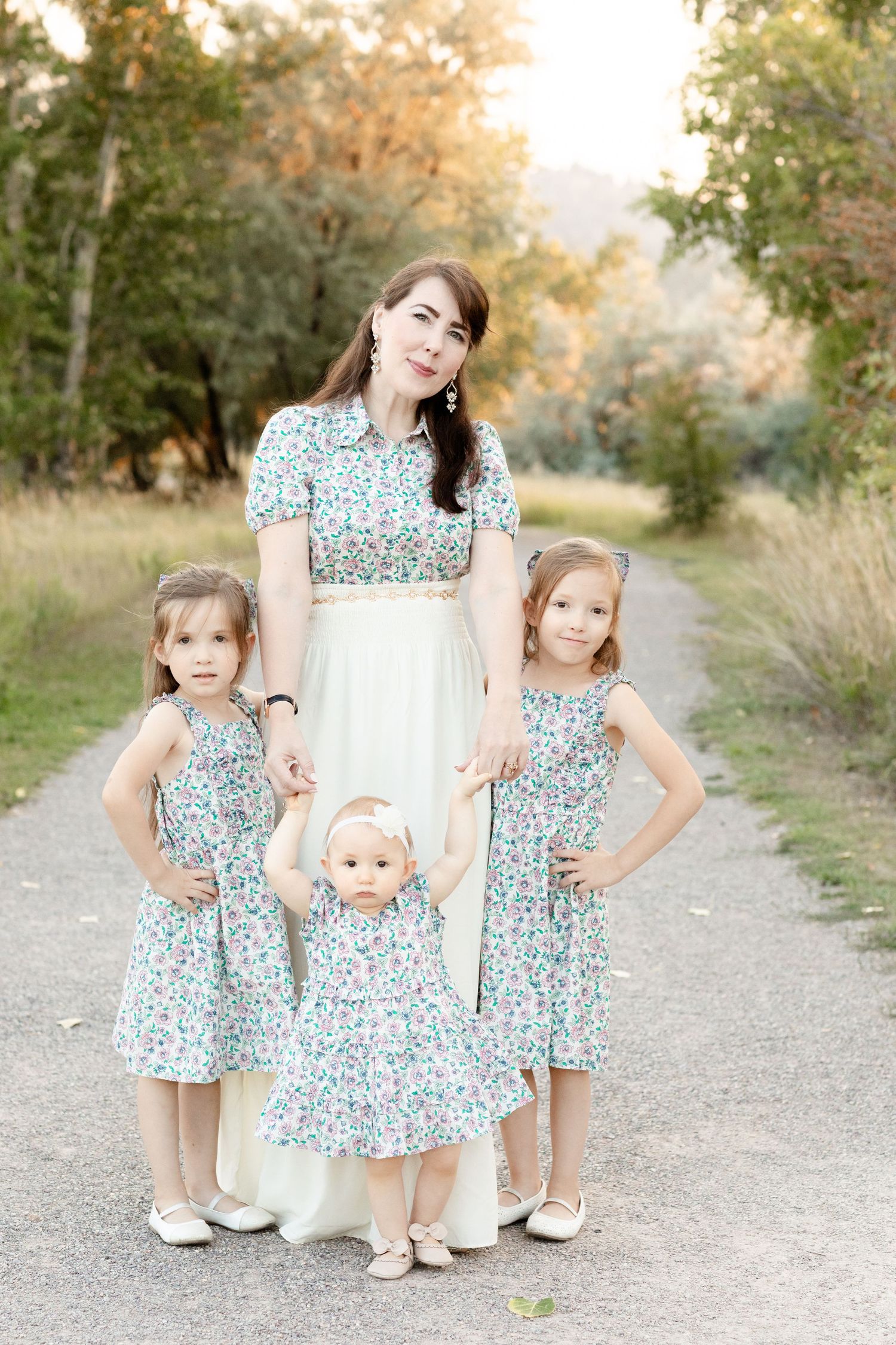 A mother in a white dress poses with her three young daughters in matching floral dresses outdoors.