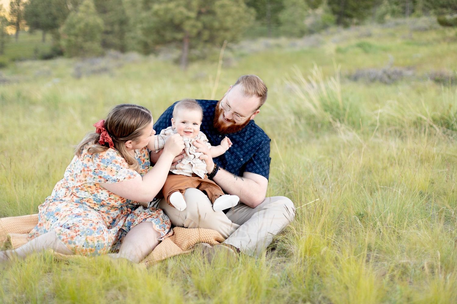 Parents playing with baby sitting in lush green grass during outdoor family photo session.