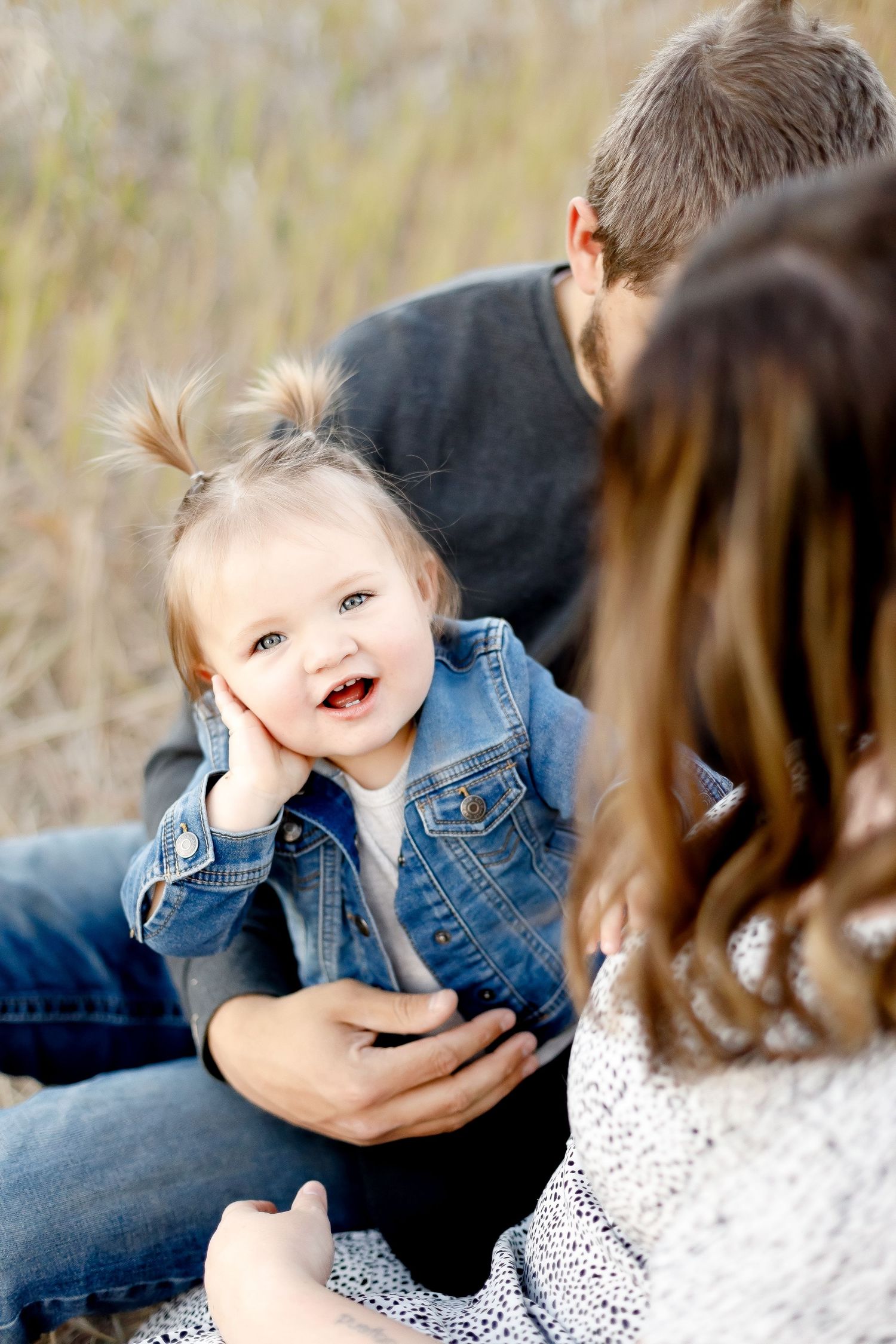 Cheerful toddler girl in denim jacket rests her chin on her hand while sitting with her parents.