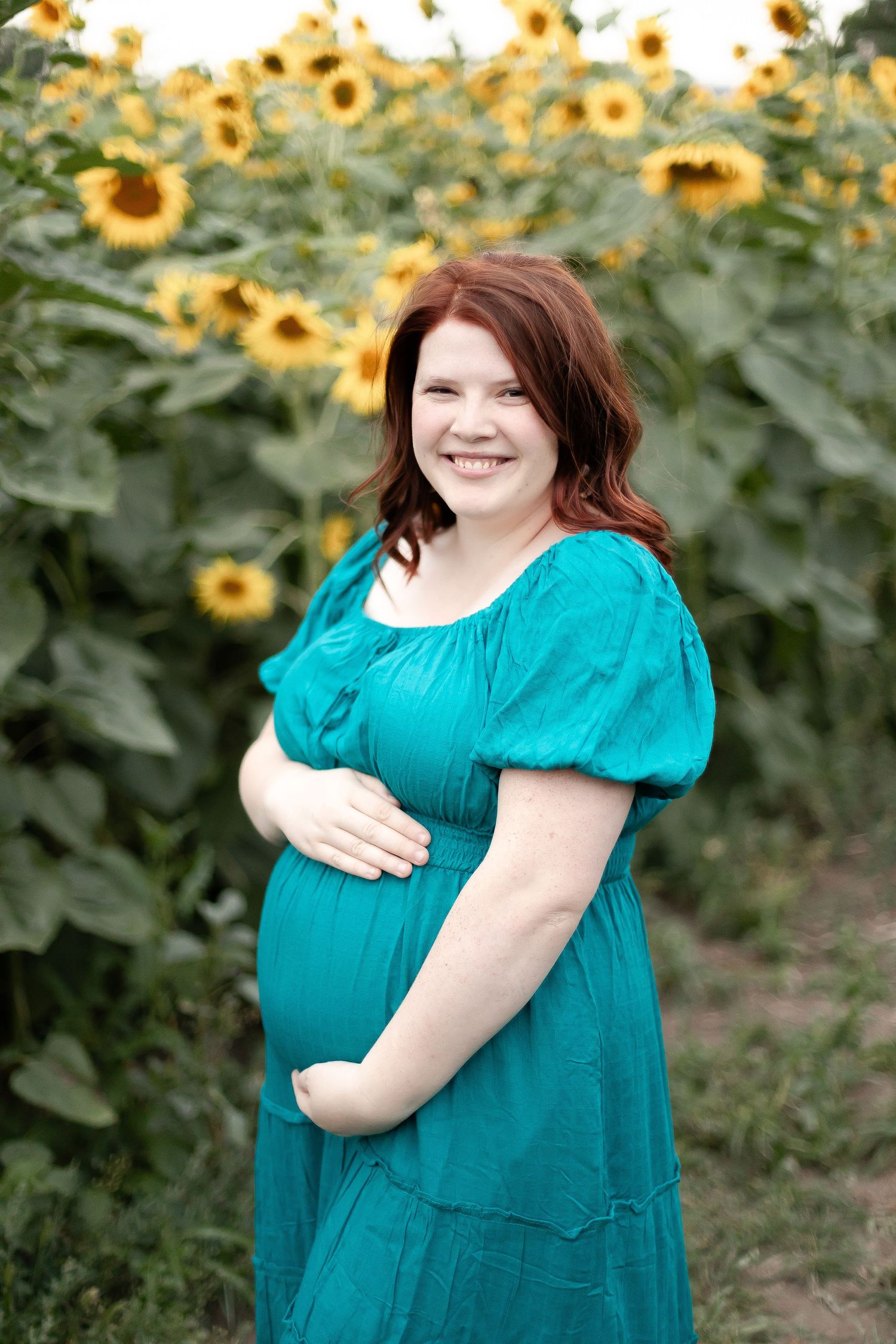 Smiling pregnant woman in teal dress holds baby bump while posing in a bright sunflower field.