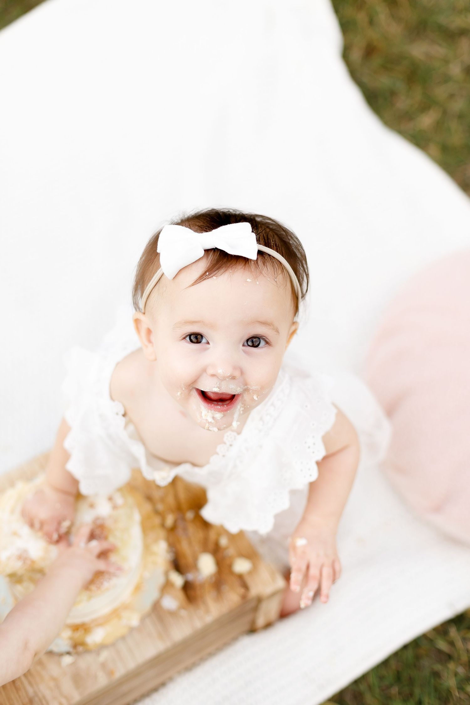 Happy baby girl in white lace dress with bow headband laughing while smashing first birthday cake outdoors.