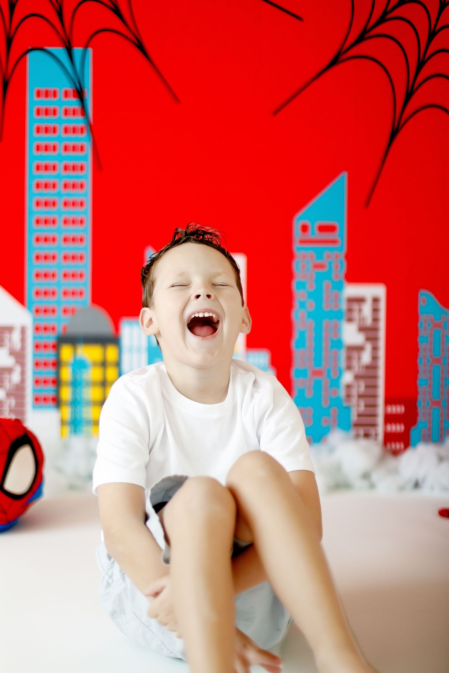 A laughing boy sits in front of a Spiderman-themed red backdrop with cityscape decorations.