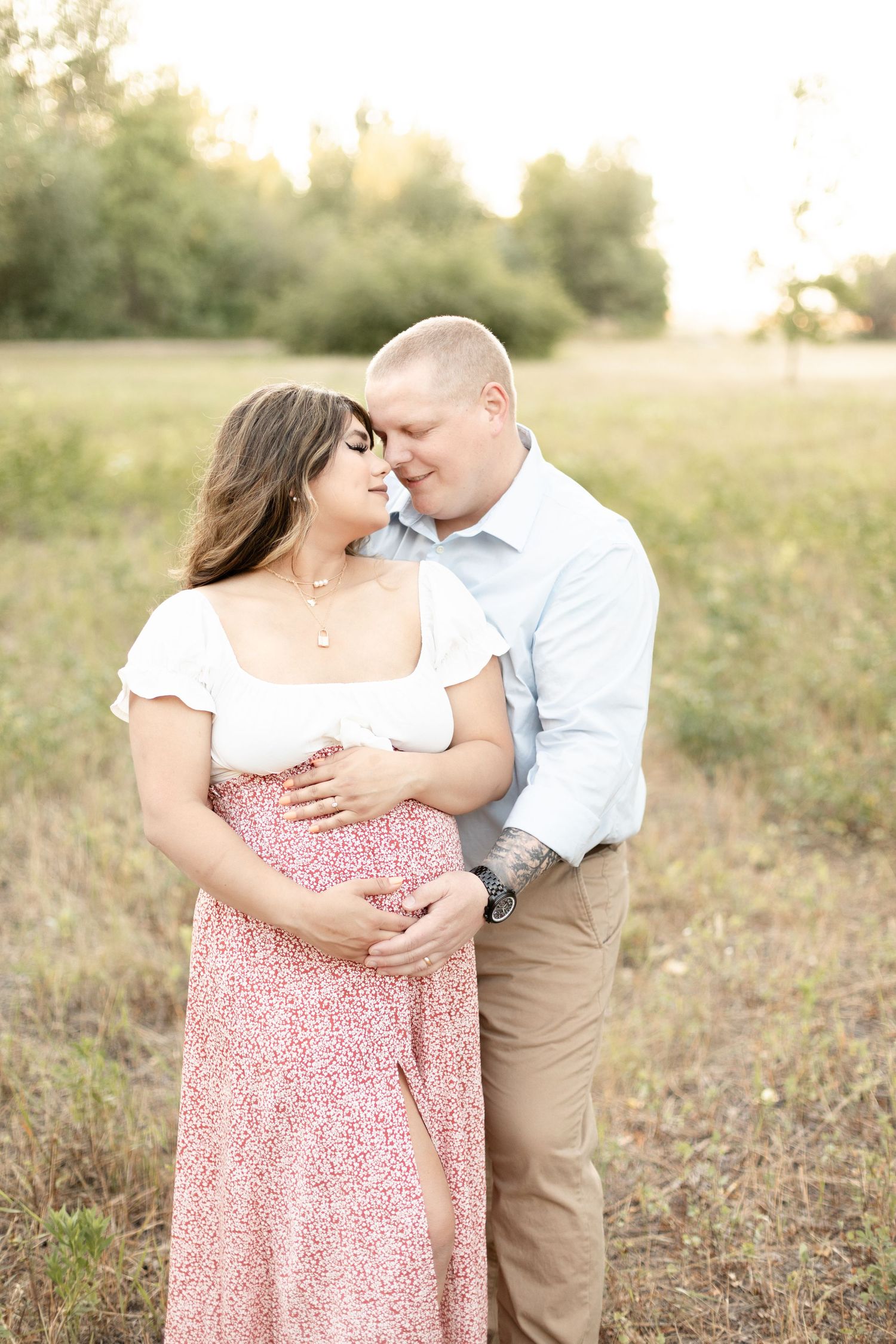 Couple embracing in golden field, pregnant woman in white and floral dress with partner during outdoor maternity session.