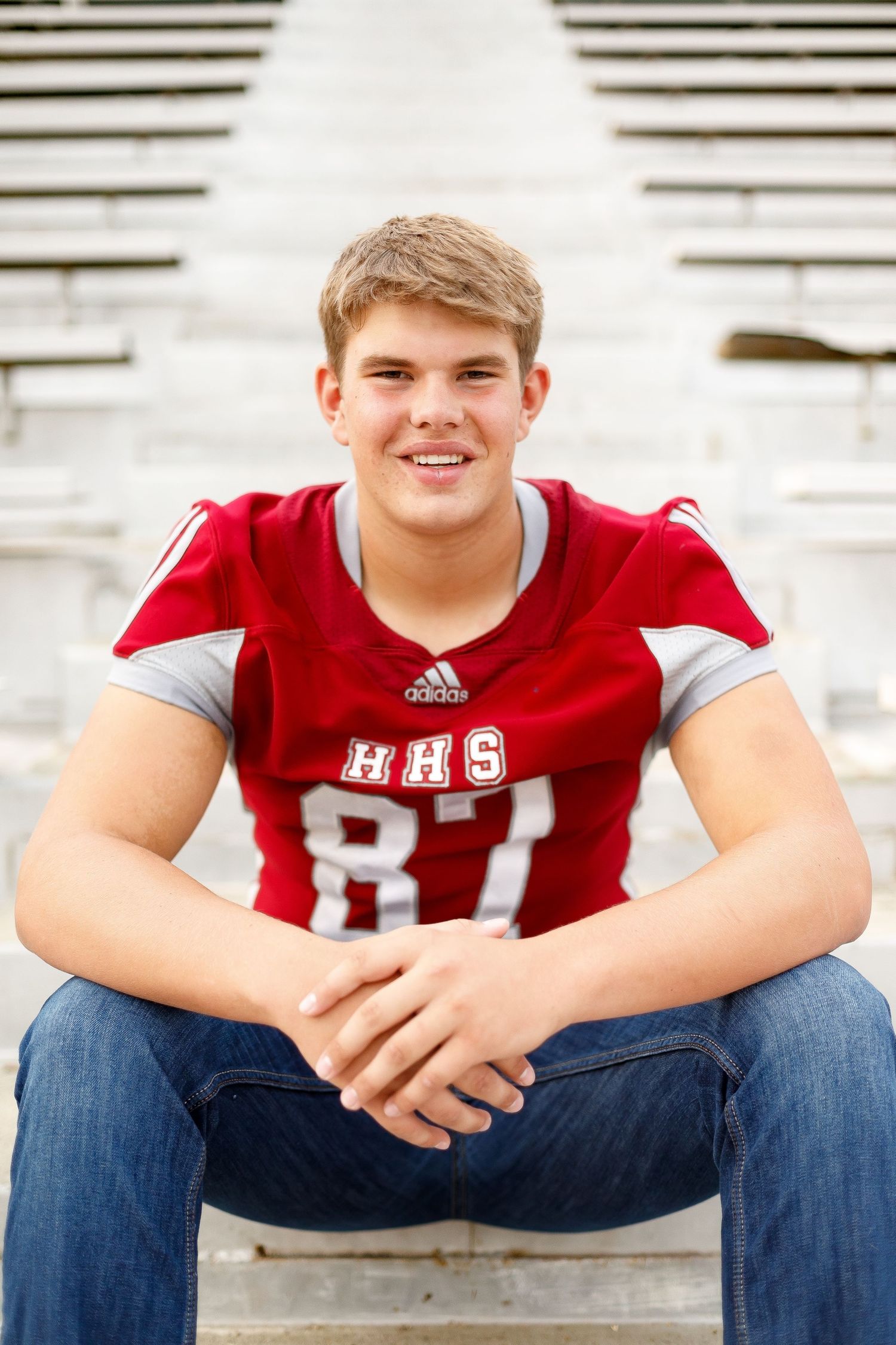 Smiling teen in HHS #87 red football jersey sitting on bleachers with jeans and white shoes.