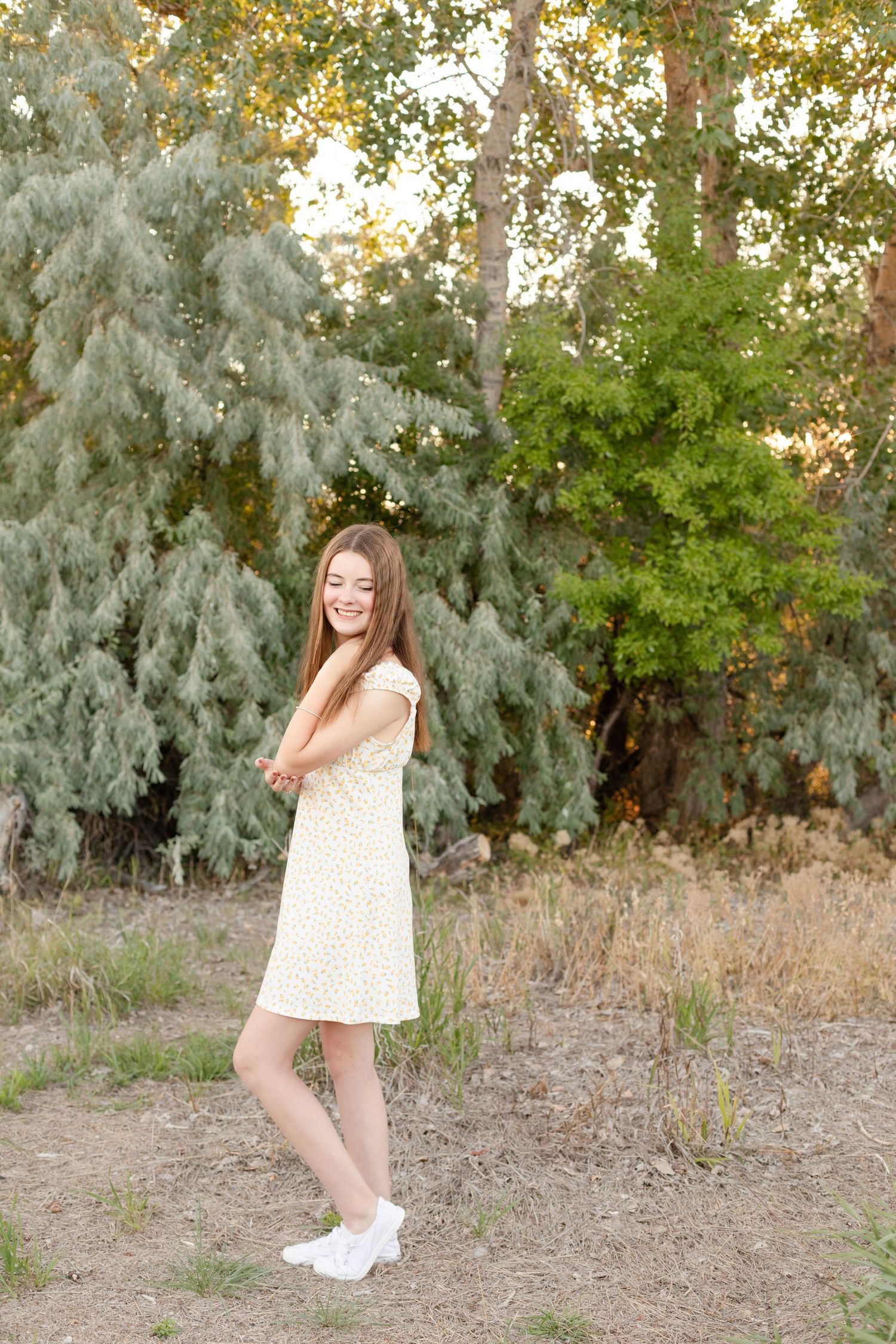 Happy teenage girl in yellow floral dress laughing outdoors with lush green pine trees behind her.