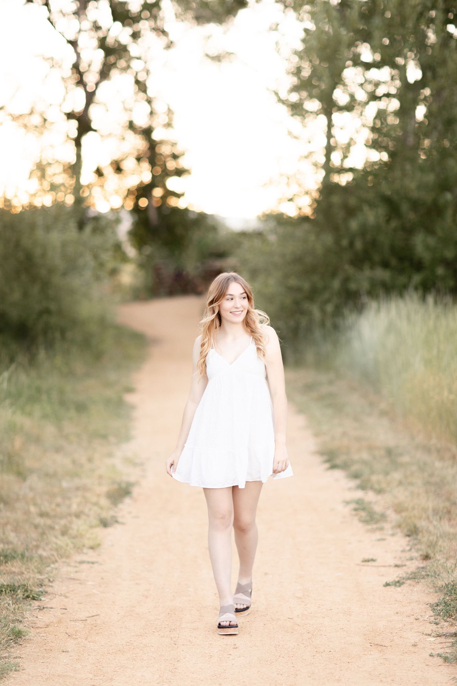 Young woman in white sundress walking on dirt path surrounded by green trees in warm golden light.