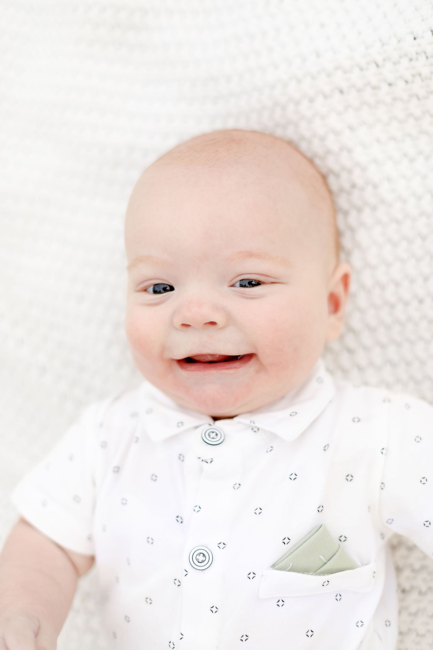 Adorable smiling baby in white outfit with pocket square lying on white blanket.