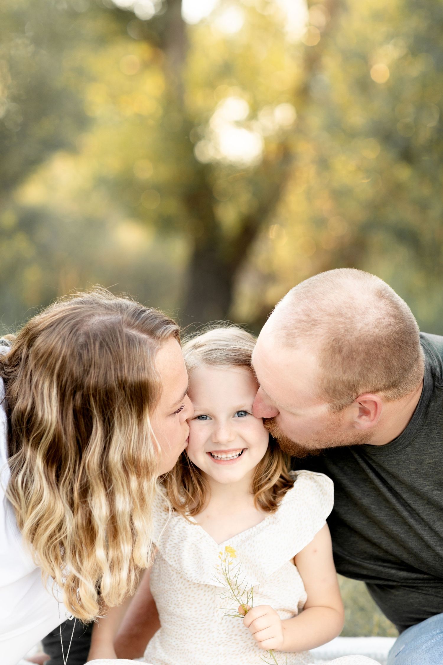 Parents kissing their smiling young daughter on the cheeks outdoors in a warm, sunlit park setting.