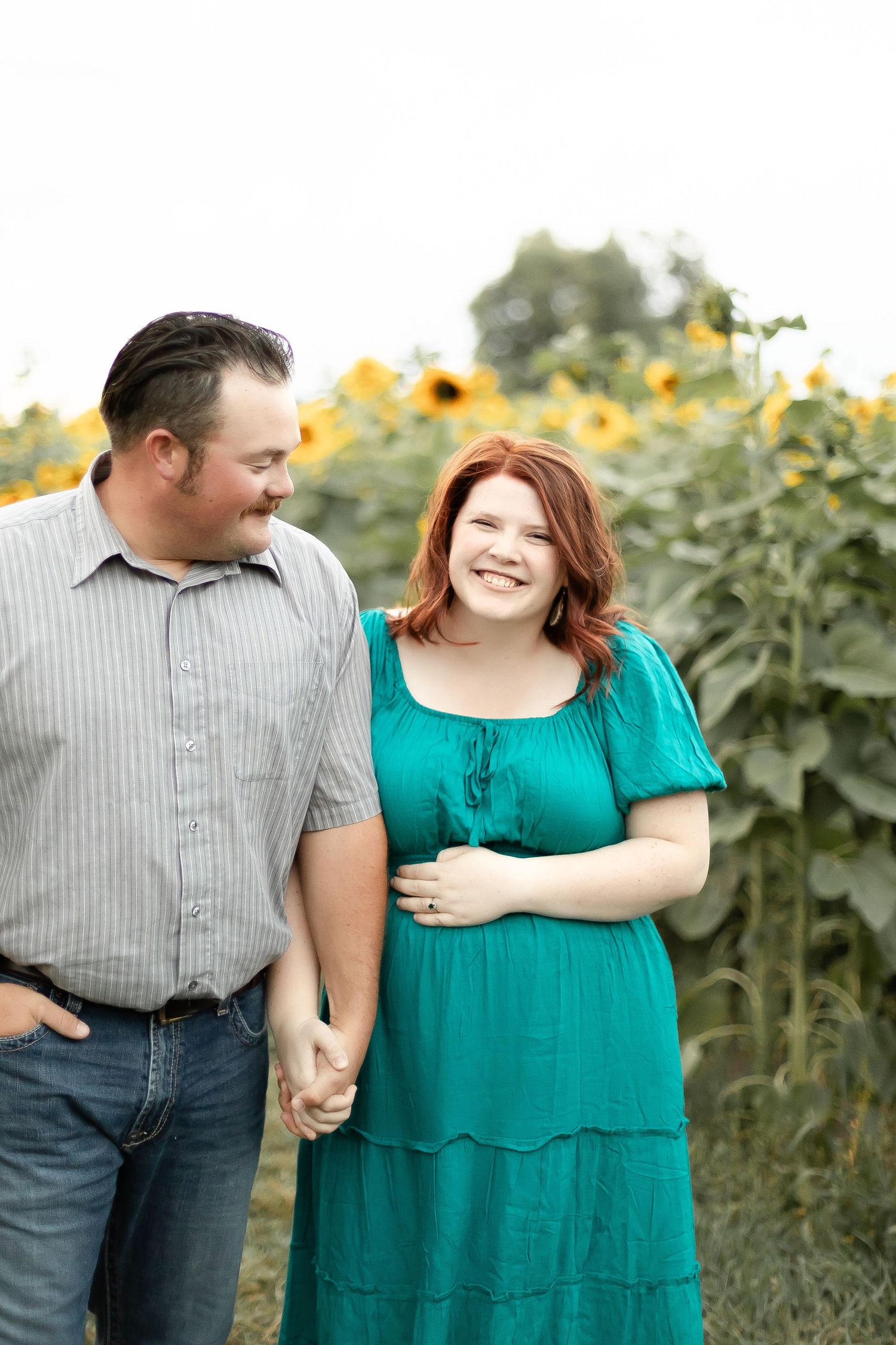Laughing pregnant woman in teal dress walks hand-in-hand with partner along a sunflower field path.