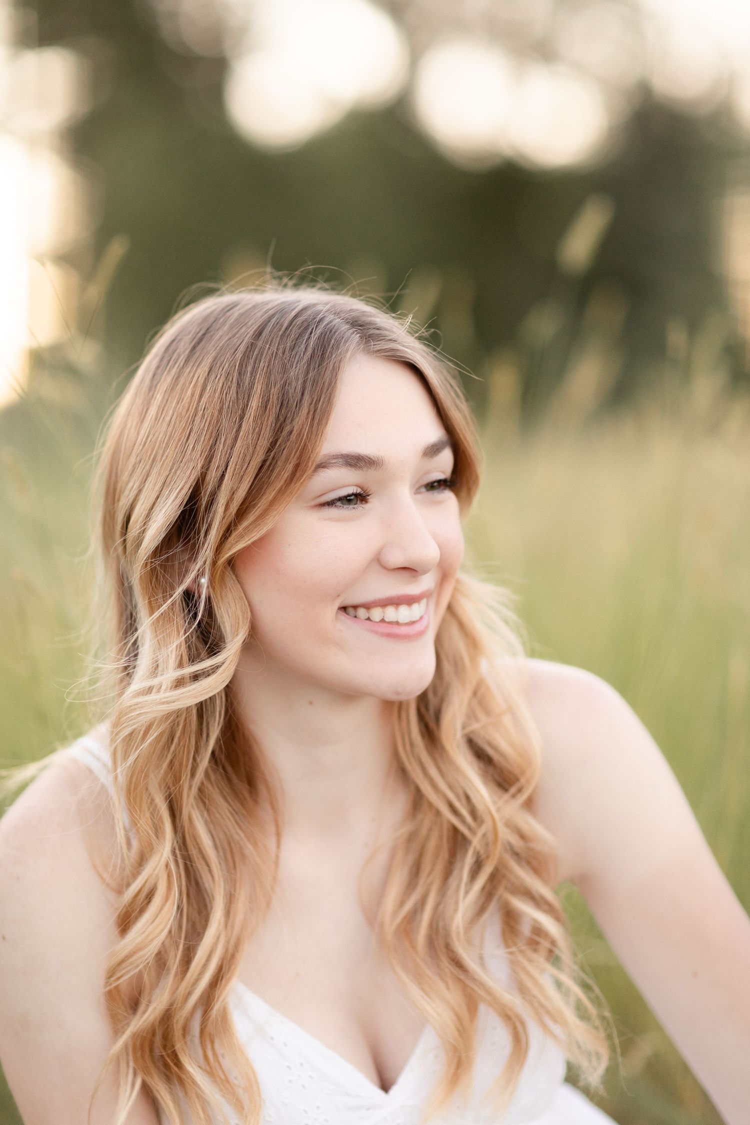 Young woman with long blonde wavy hair smiling outdoors in a field, wearing a white dress in soft golden light.