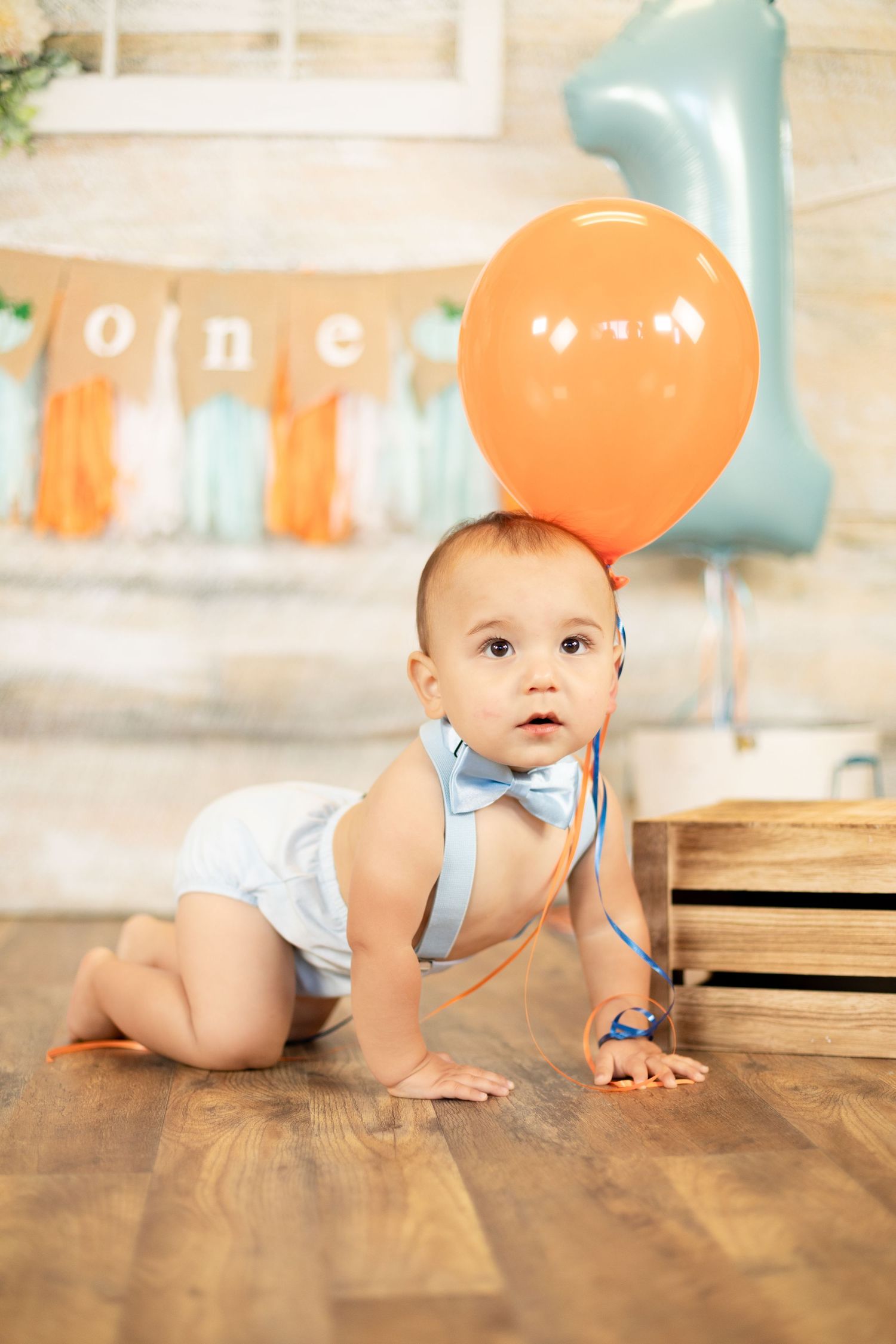 Baby boy in white diaper and blue bow tie crawling with orange balloon tied to him at first birthday party.