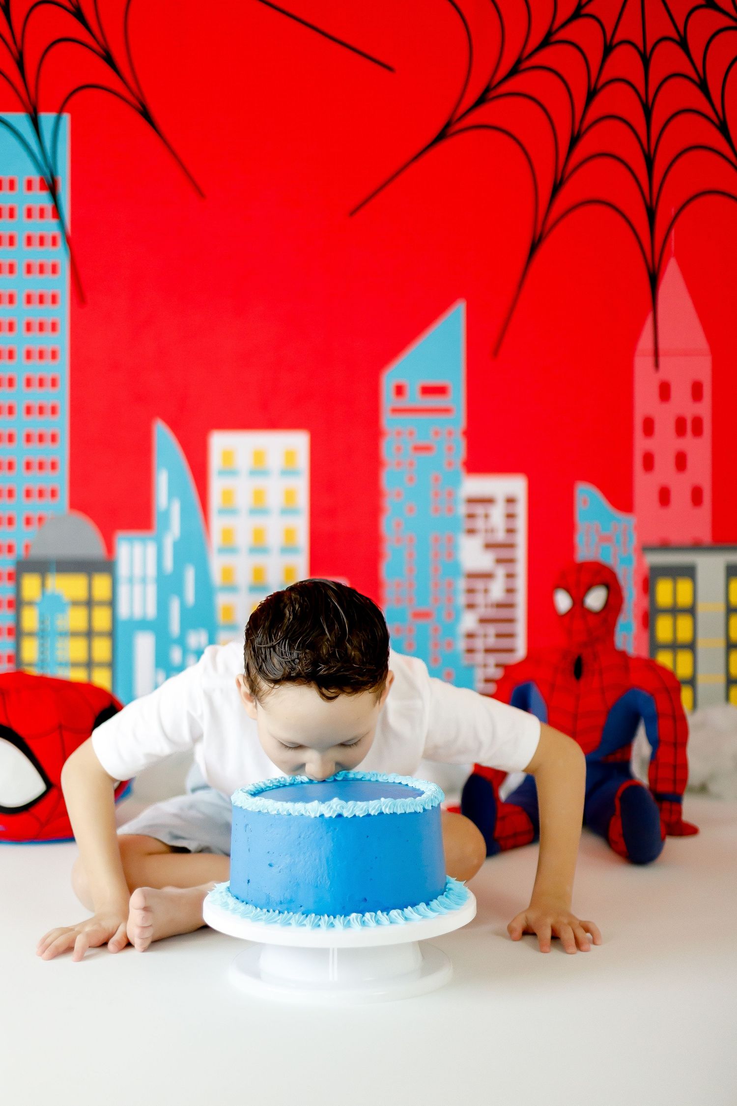 A boy smashes his face into a blue birthday cake at a Spiderman-themed party.
