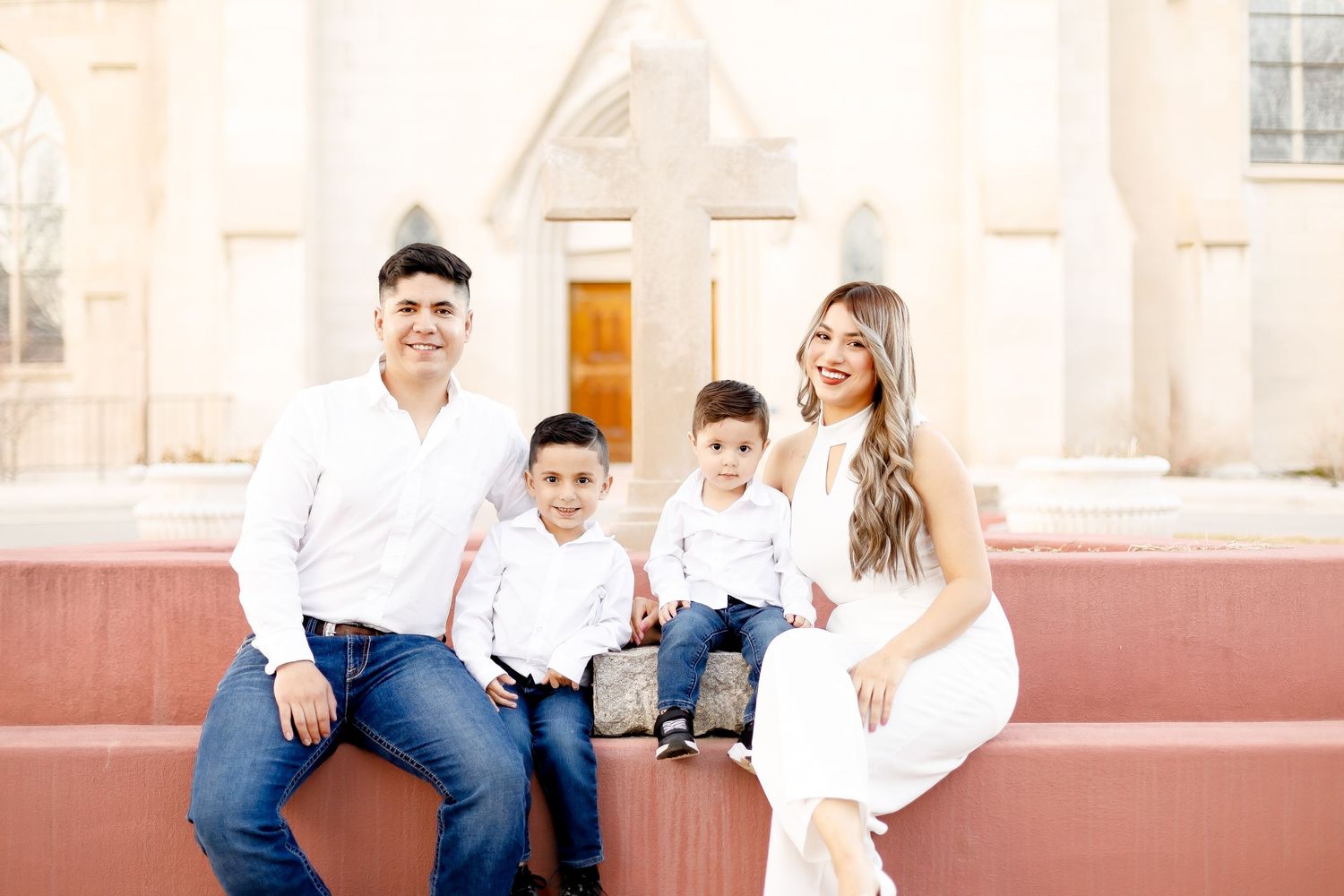 Happy family of four in white outfits sitting on steps in front of a church.