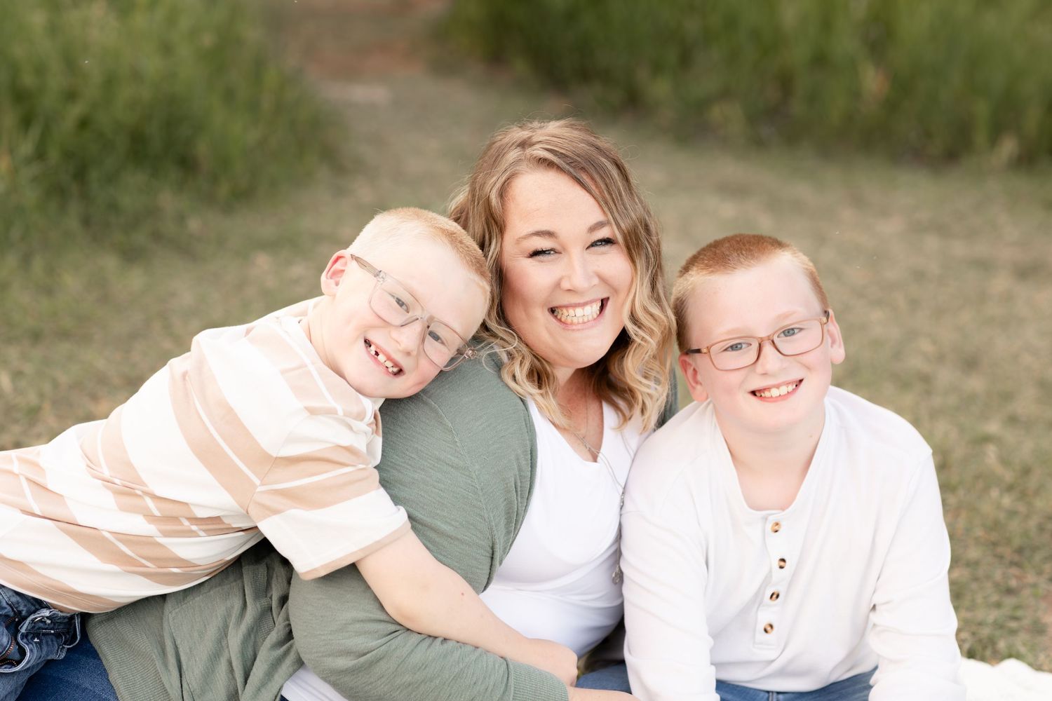 A smiling mother hugs her two red-haired sons wearing glasses in a green outdoor field.