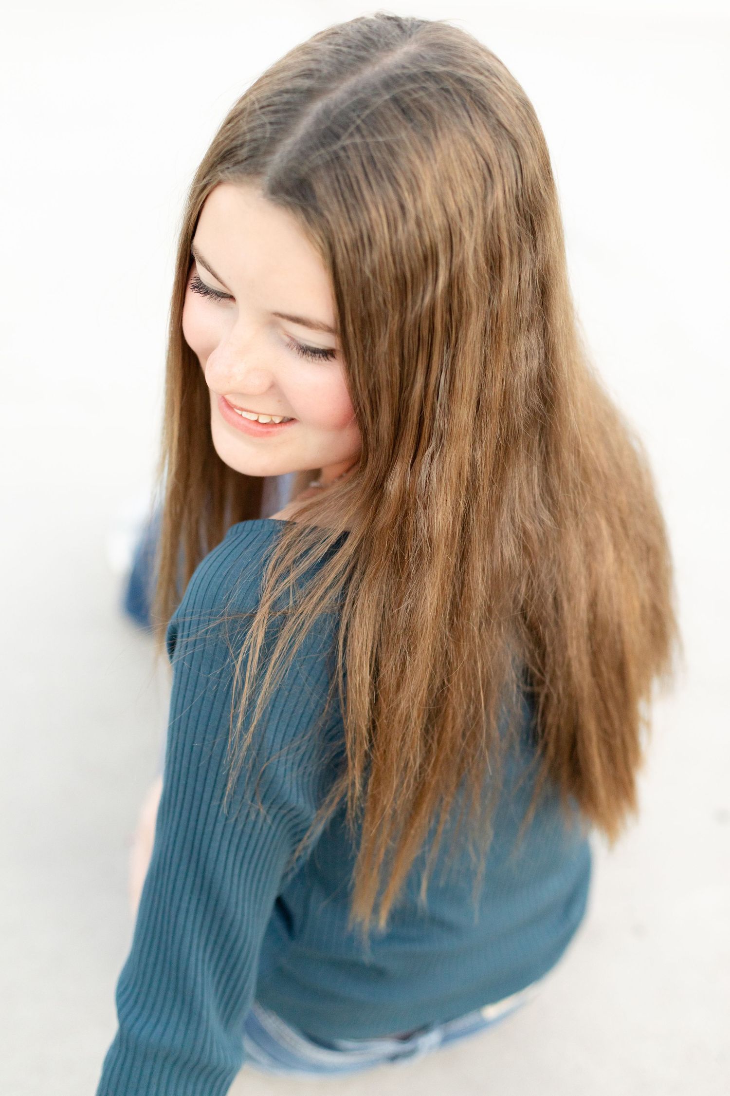 Teenage girl with long brown hair smiling downward wearing a teal ribbed top against white background.