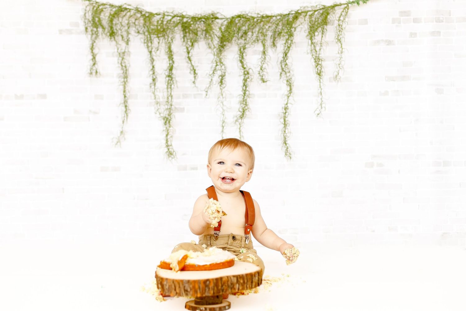 A cute baby boy in suspenders smashing his first birthday cake on a rustic wooden stump backdrop.
