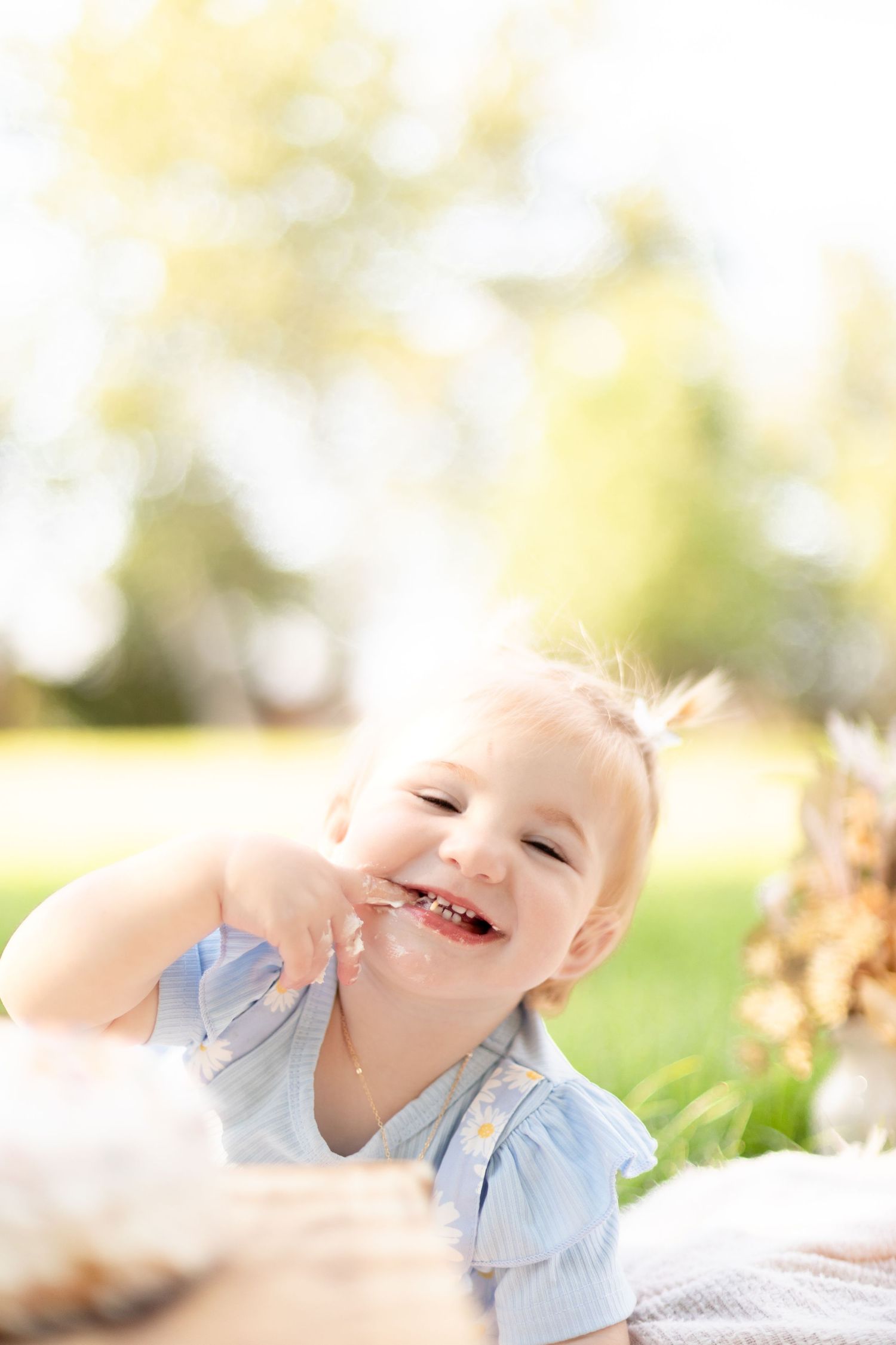 Laughing baby girl with cake on her face enjoying her first birthday smash cake outdoors in a sunny park.