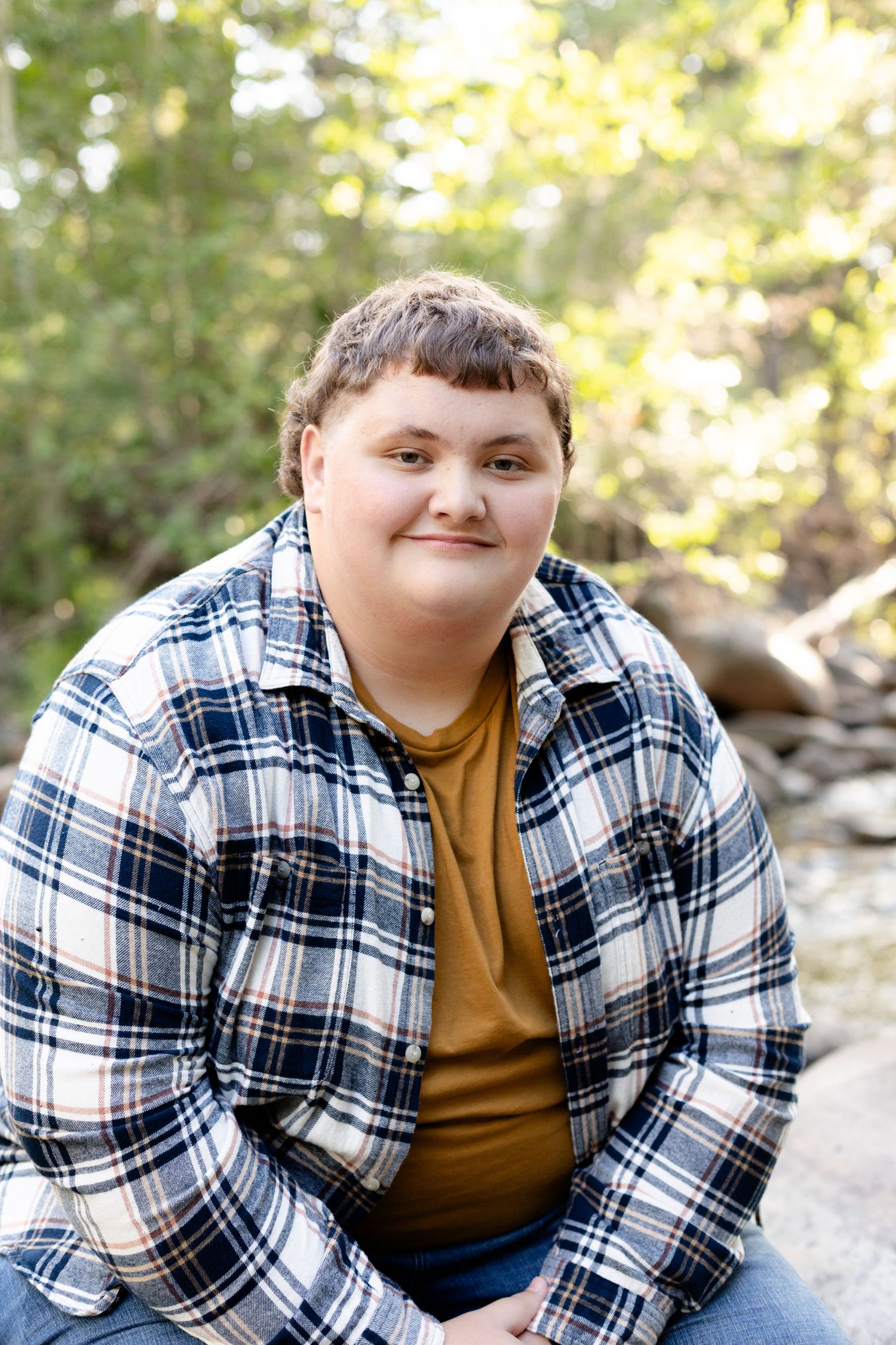 Young person in plaid flannel shirt smiling softly while seated on rocks in a sunlit forest setting.