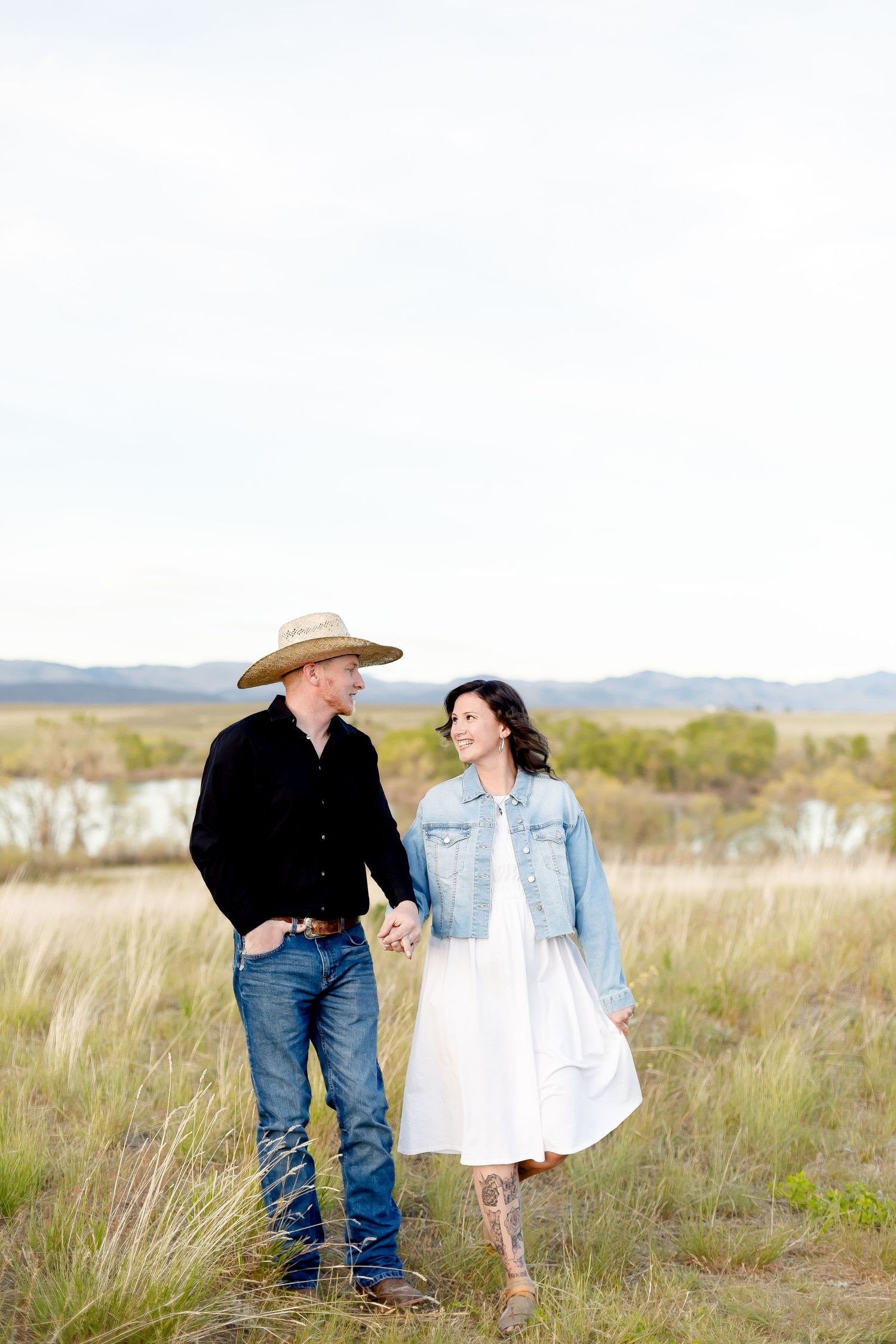 Country couple holding hands walking through a grassy field, man wearing cowboy hat and woman in white dress.
